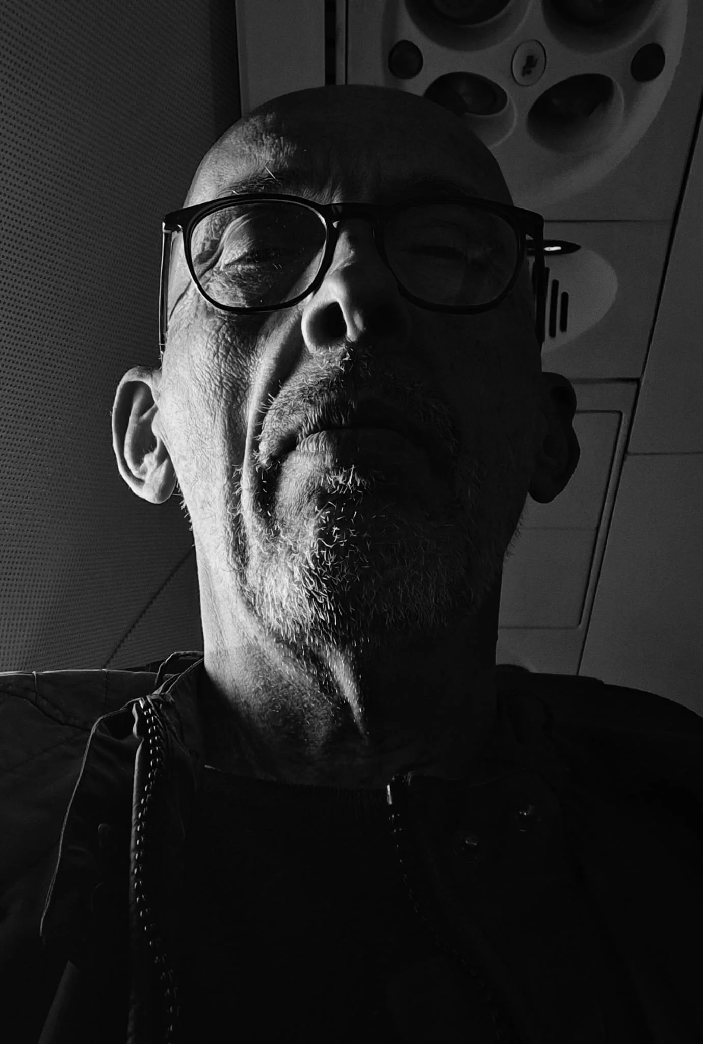 Ritratto in bianco e nero di un uomo con occhiali e barba, vestito in nero, con sfondo di un soffitto con luci, scattato in un ambiente interno.