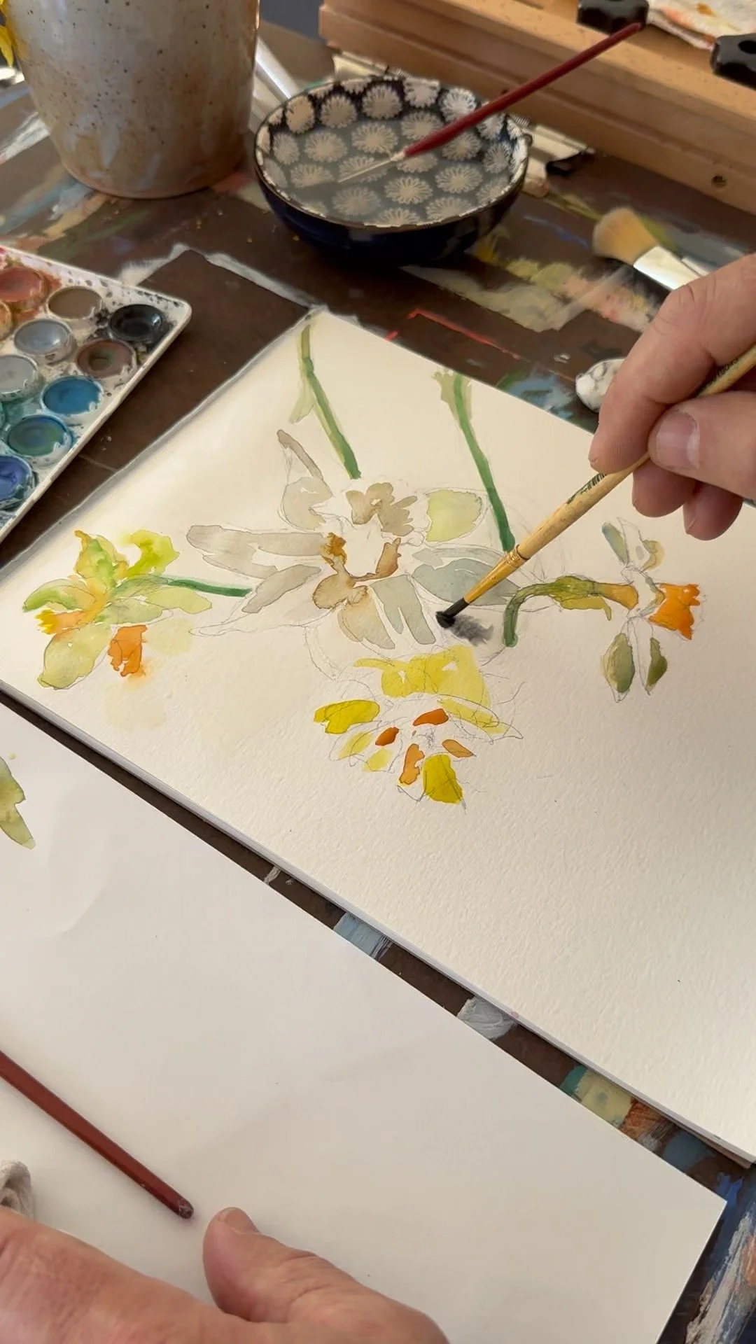 A person painting a floral watercolor artwork on paper, with various watercolor paints and brushes on the table.