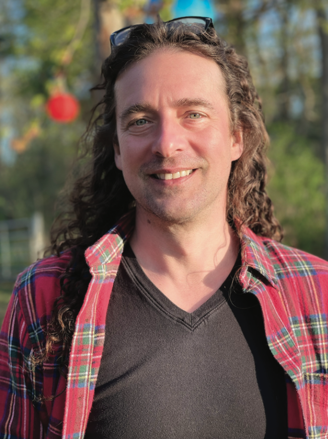 Smiling man with long curly hair wearing a black t-shirt and a red plaid shirt, sunglasses on his head, outdoors with blurred trees and a red ornament in the background.