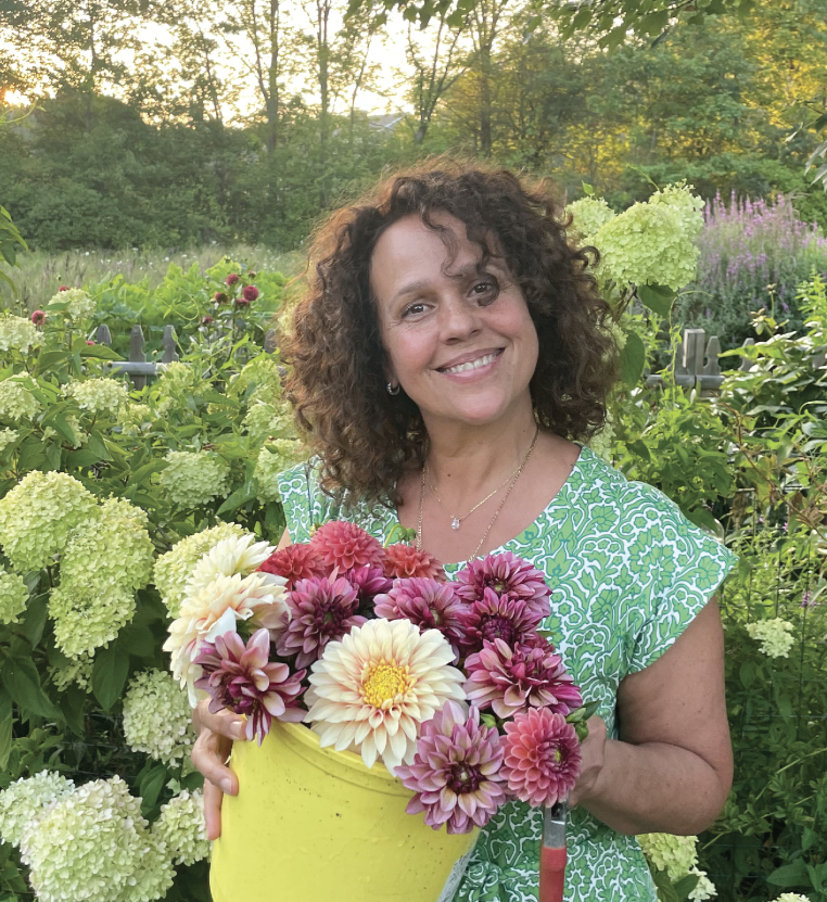 A woman with curly hair smiling and holding a yellow pot filled with pink, purple, and white dahlias in a garden with lush greenery and trees in the background.