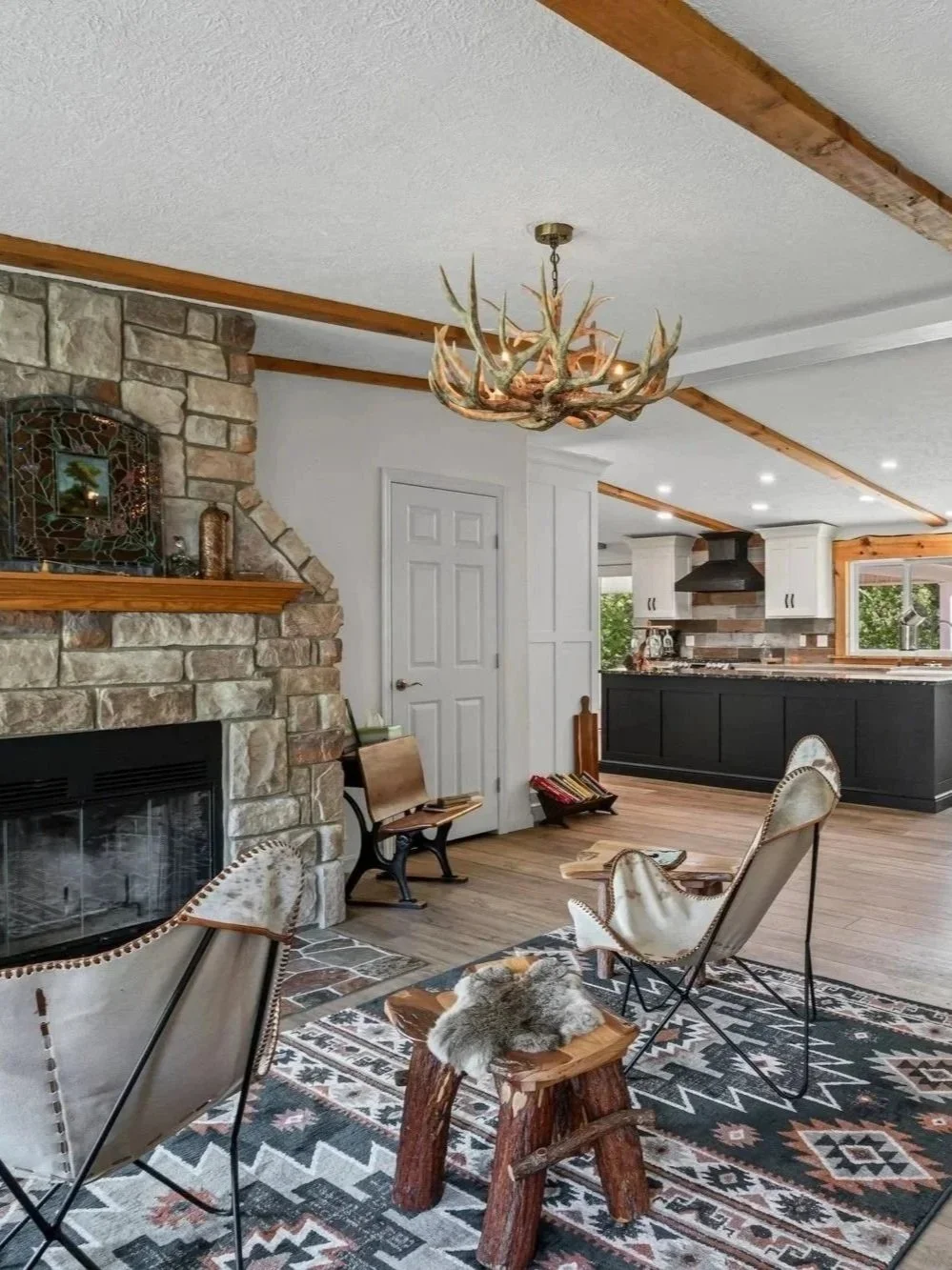 Living room with stone fireplace, wooden beams, rustic chandelier, leather lounge chairs, animal hide rug, and views of the kitchen with black cabinets.