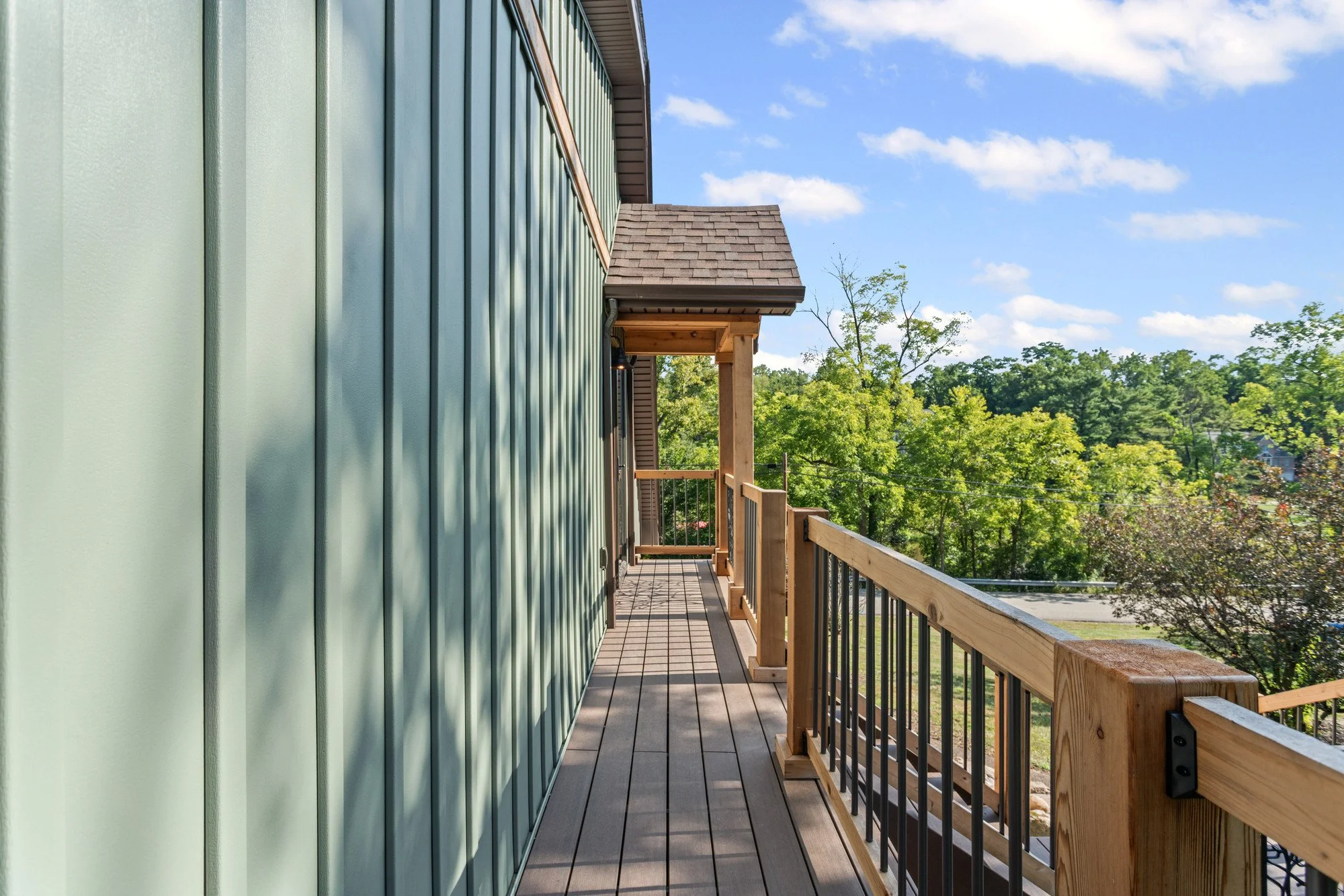 A wooden balcony with horizontal metal siding on a building, overlooking a green landscape with trees and blue sky with a few clouds.