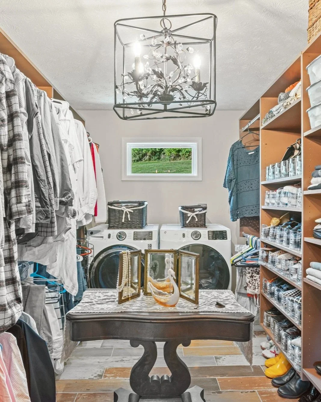 Laundry room with a chandelier, top-load washer and dryer, shelves with folded clothes and storage bins, a small window, and a decorative swan in a jewelry box on a table.