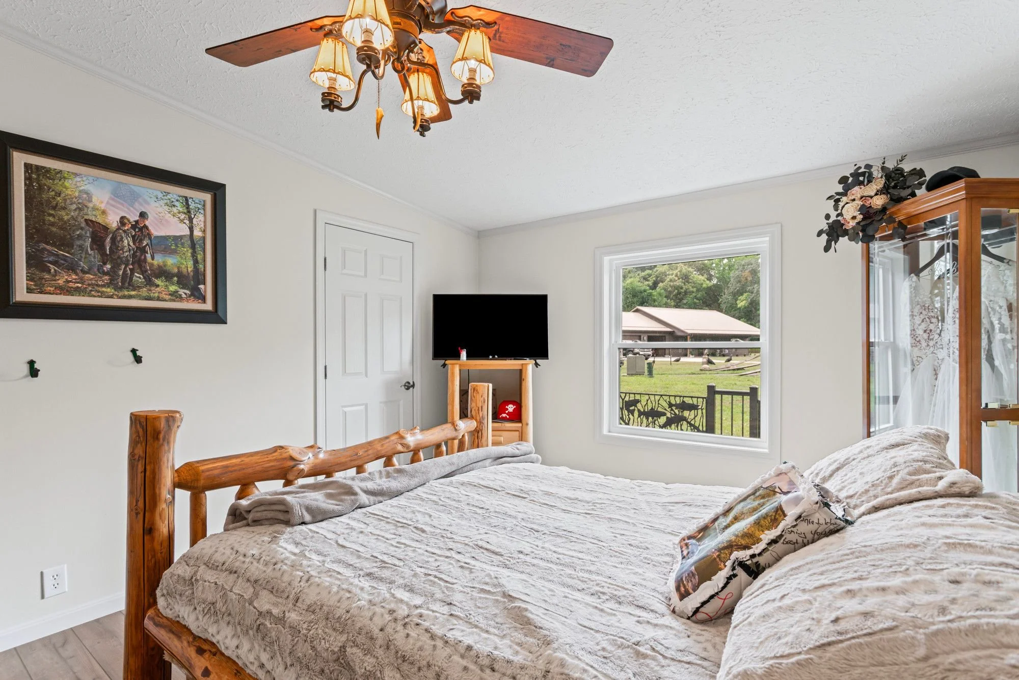 A bedroom with a wooden bed, beige blanket, and pillows. Hanging on the wall is a framed picture of hikers in a forest. There is a small, TV stand with a black television and a red item underneath. A window shows a view of green trees and a neighboring house. A wooden display cabinet with flowers on top is also in the room. A ceiling fan with lights hangs from the ceiling.