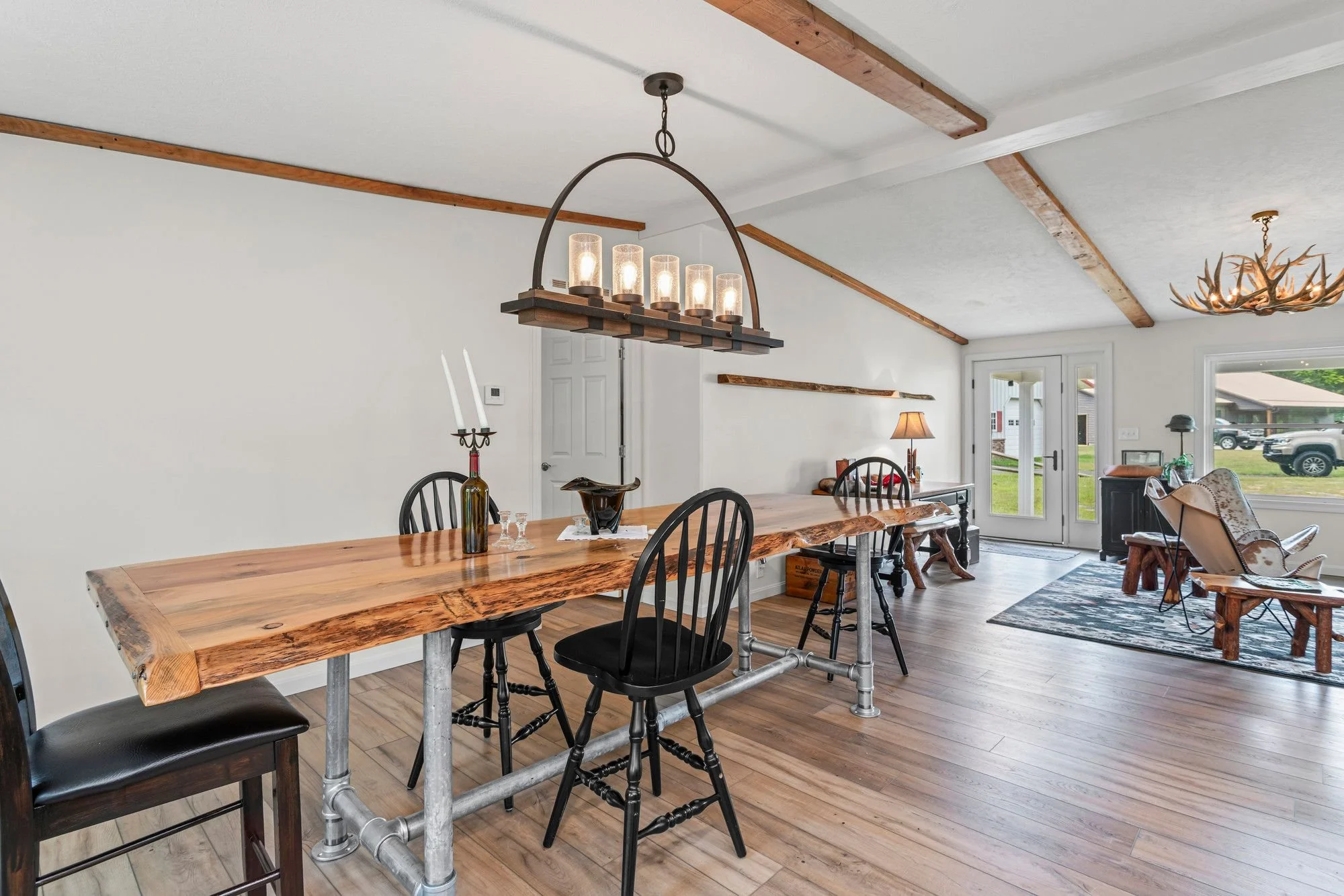 A dining room with a large wooden table, black chairs, and a chandelier with six glass candle-like lights hanging from the ceiling. The room has wooden beams on the ceiling and hardwood floors. There is a door and windows with a view of a lawn and parked cars outside.
