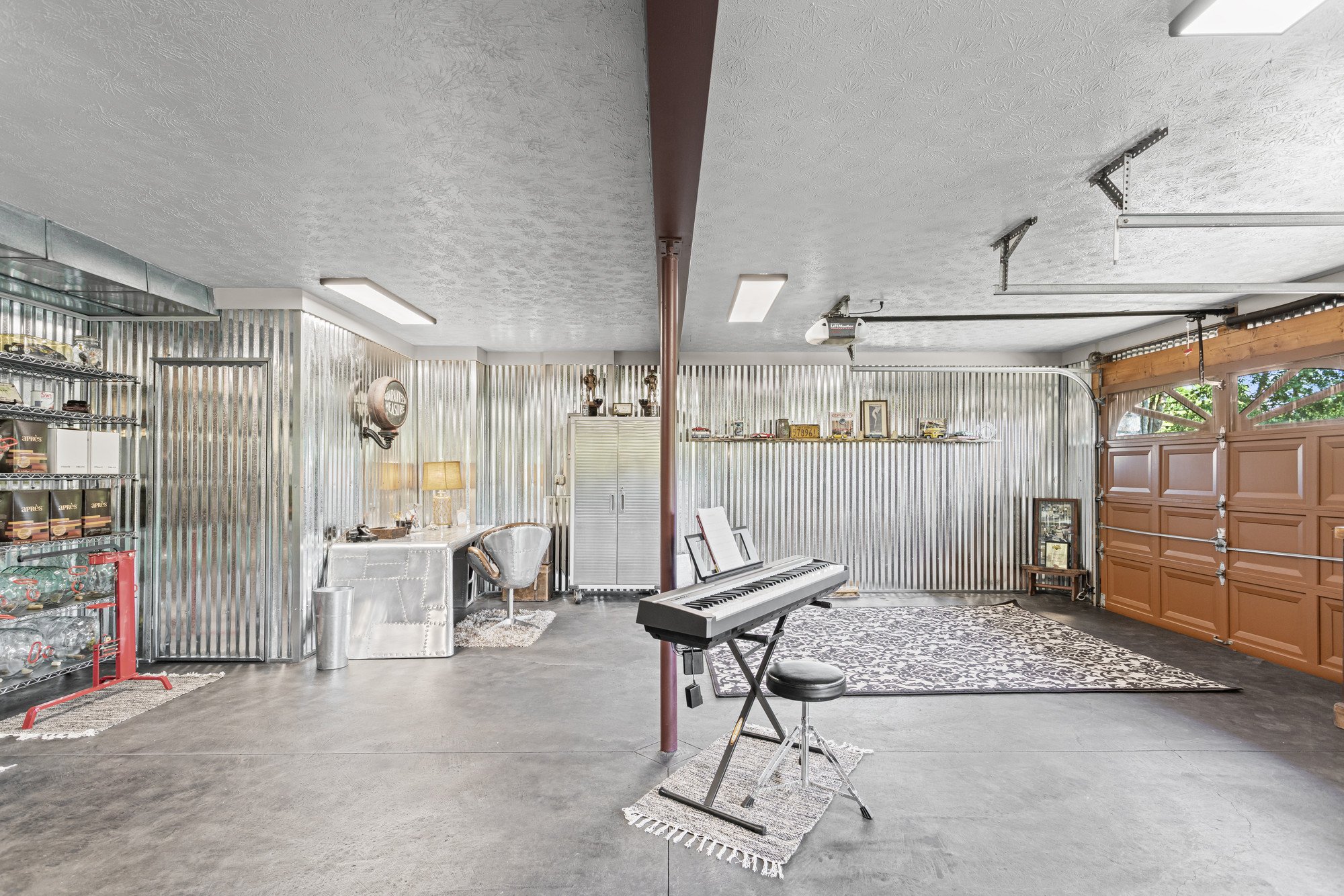 A spacious garage converted into a music studio with a digital piano, a music stand, and eclectic decor including shelves with collectibles, a vintage sign, a lamp, and various framed pictures. The garage has a corrugated metal wall, a large orange sectional garage door, and a window revealing greenery outside.
