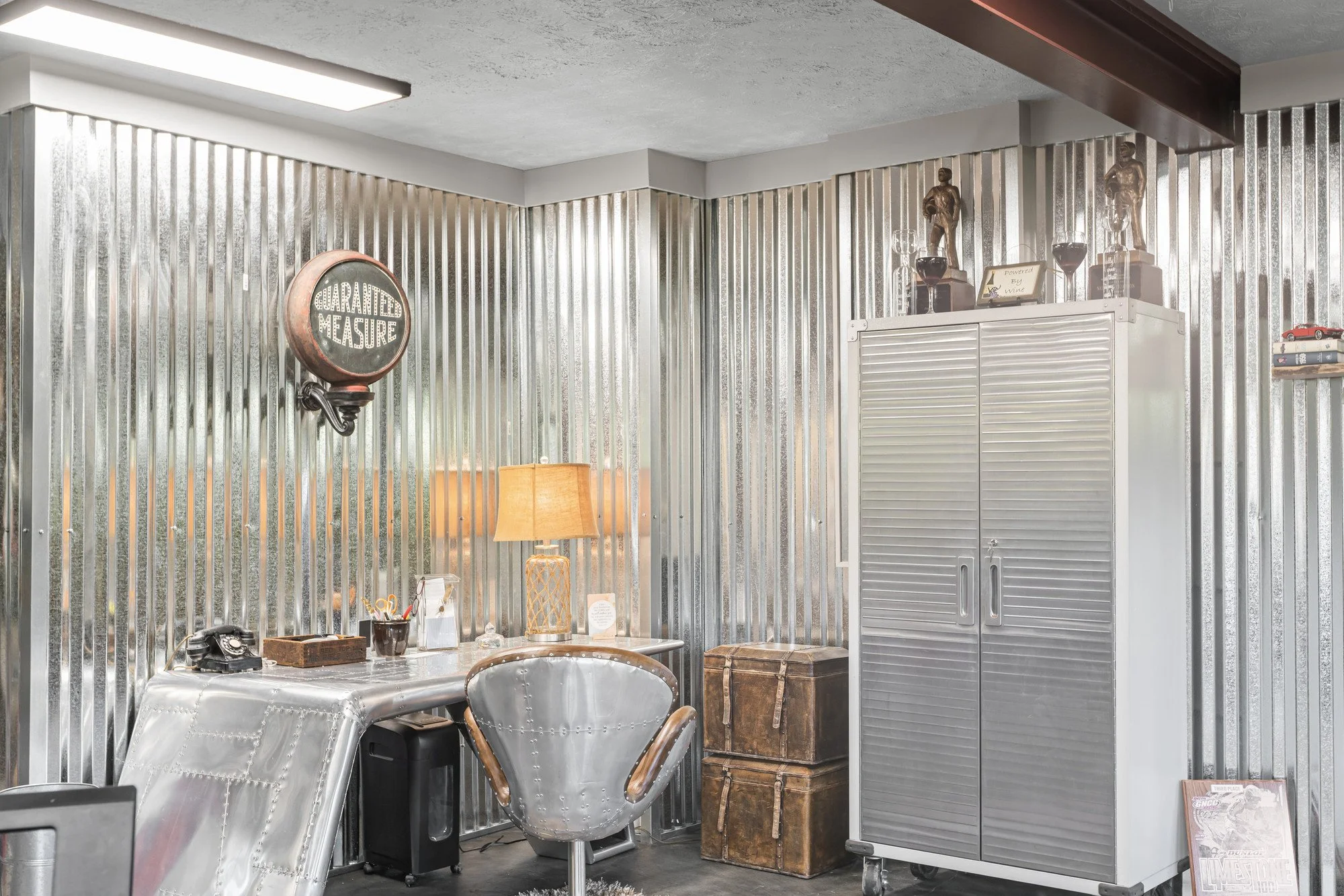 Interior of a room with corrugated metal walls and ceiling, featuring a sign that reads 'Guaranteed Measitre', a table with various items including a vintage wall phone, lamp, and small decor, as well as a metal chair, a gray cabinet with trophies and wine glasses on top, and stacked vintage trunks.