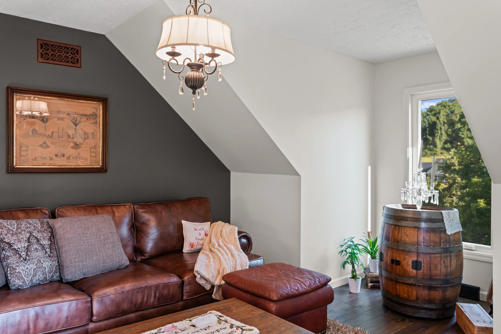 Living room with a leather sofa, decorative pillow, fringe blanket, and a framed picture on the dark wall. A chandeliers hangs from the ceiling. There is a window with a scenic view and a table with a chandelier made of glass in the corner.