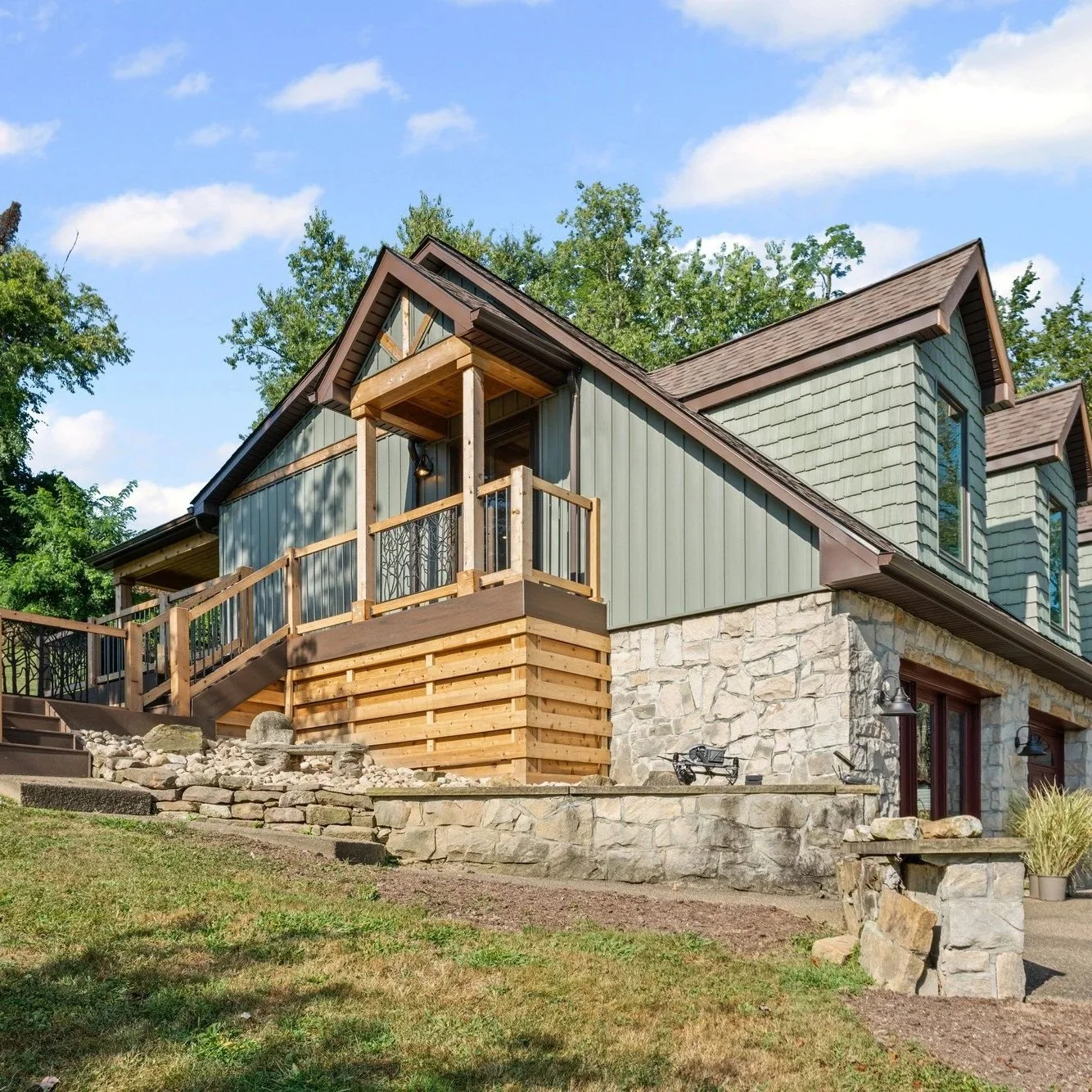 A two-story house with green siding, stone foundation, and a wooden balcony on the second floor, surrounded by trees and a grassy yard under a blue sky.