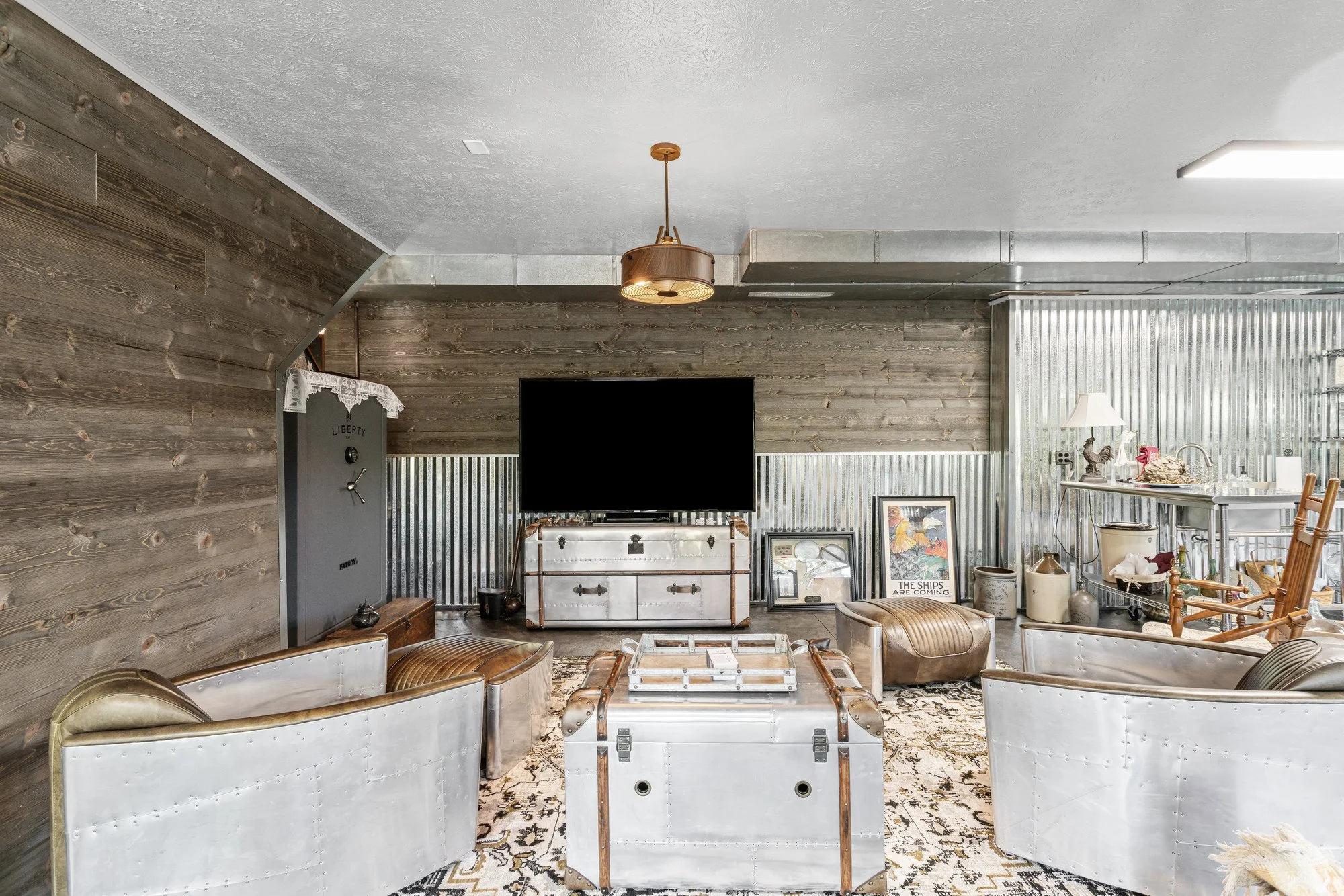 Living room with rustic wooden wall and ceiling, vintage metal trunk coffee table, leather armchairs, framed artwork, and mix of modern and vintage decor, including a lamp and various small objects.