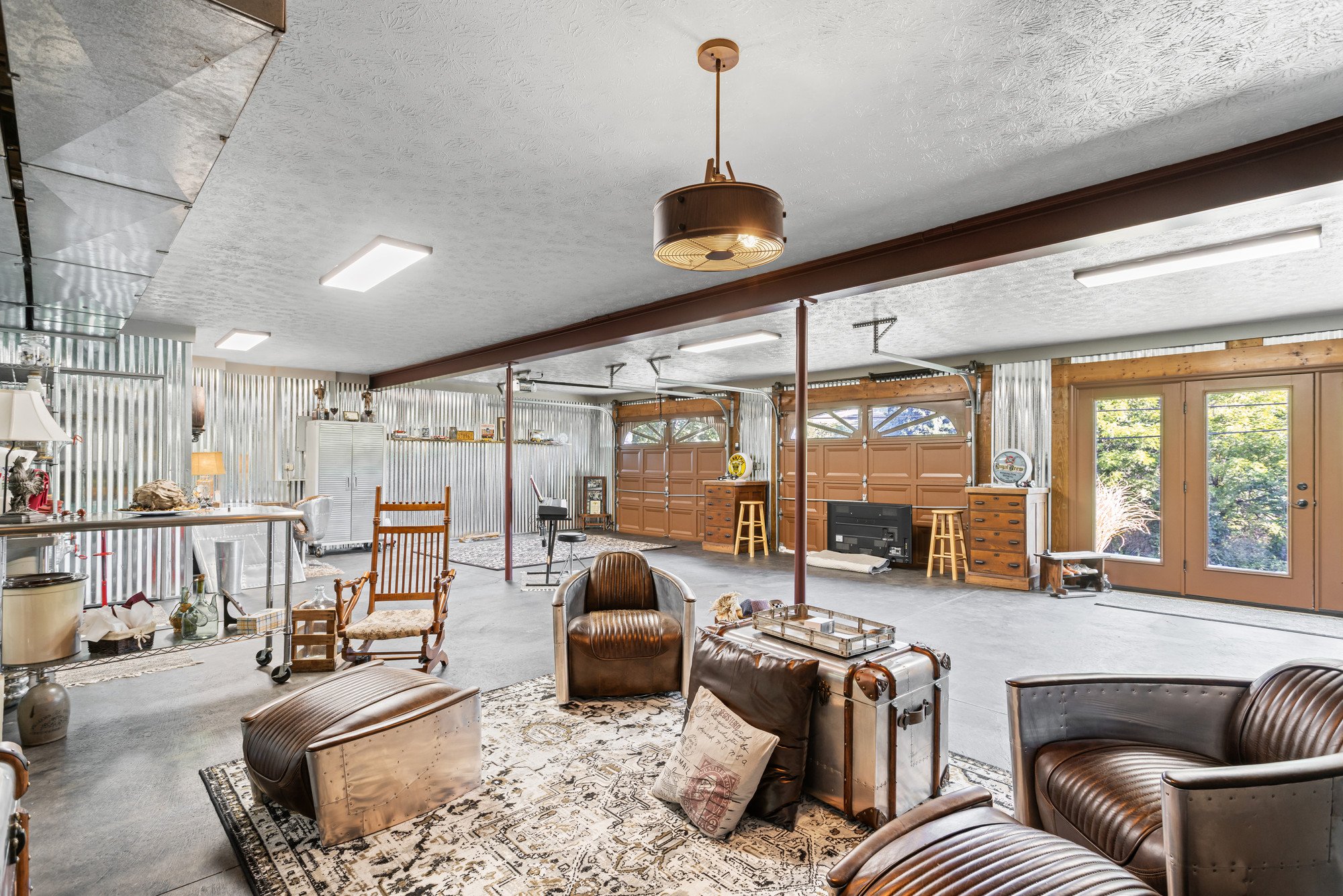 Interior of a spacious garage converted into a living space, featuring leather furniture, wooden chairs, a television, and decorative items, with metal and wood wall panels and large garage doors.