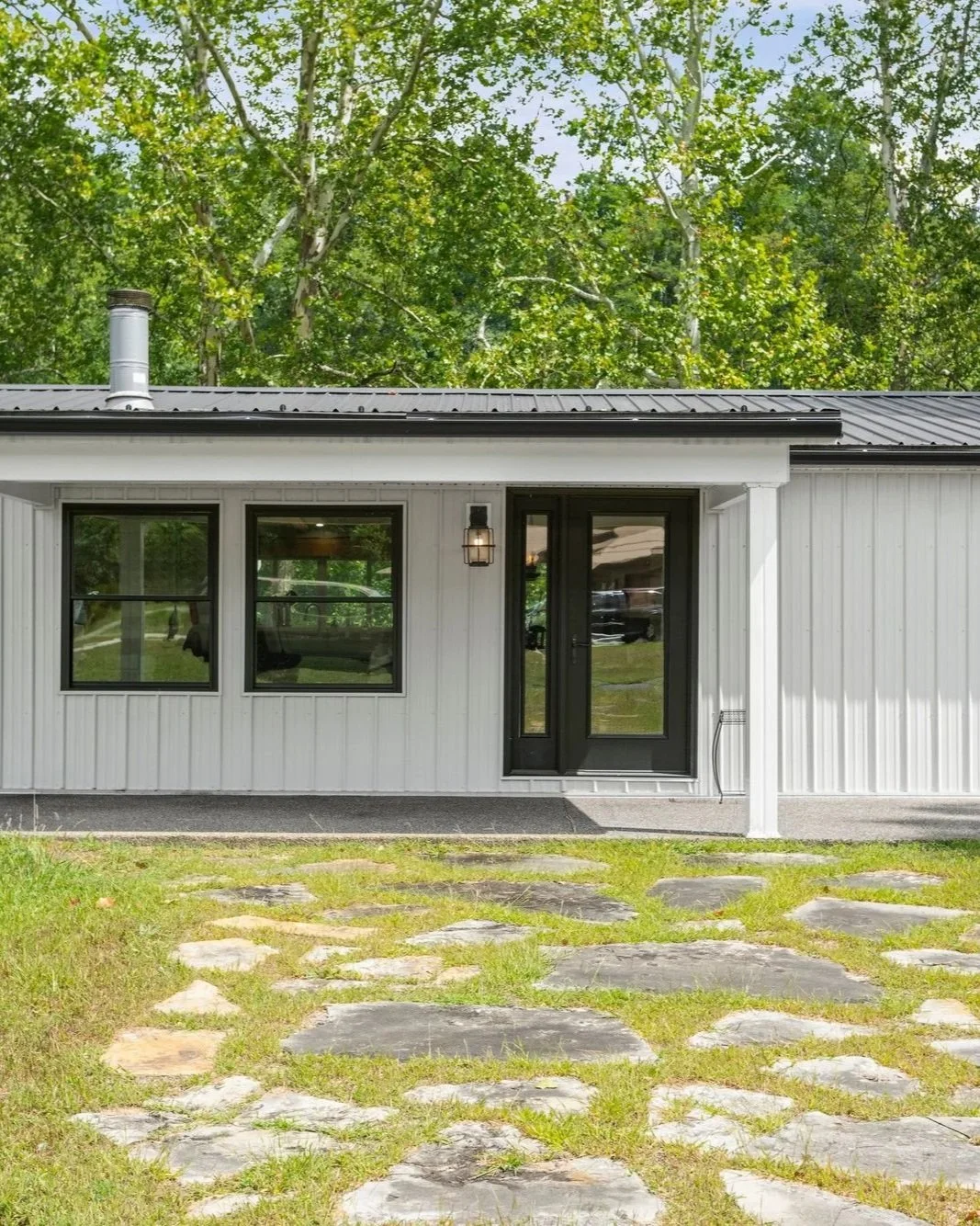 Front view of a modern house with white vertical siding, black-framed windows and door, gray metal roof, outdoor wall lantern, stone pathway, and green trees in the background.