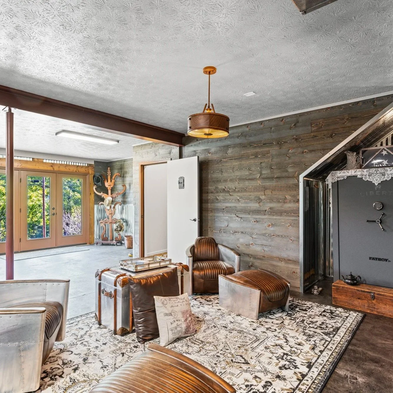 Interior living room area with a wooden wall, leather armchair, vintage trunk, and patterned rug. Glass doors lead to outdoor space with greenery.