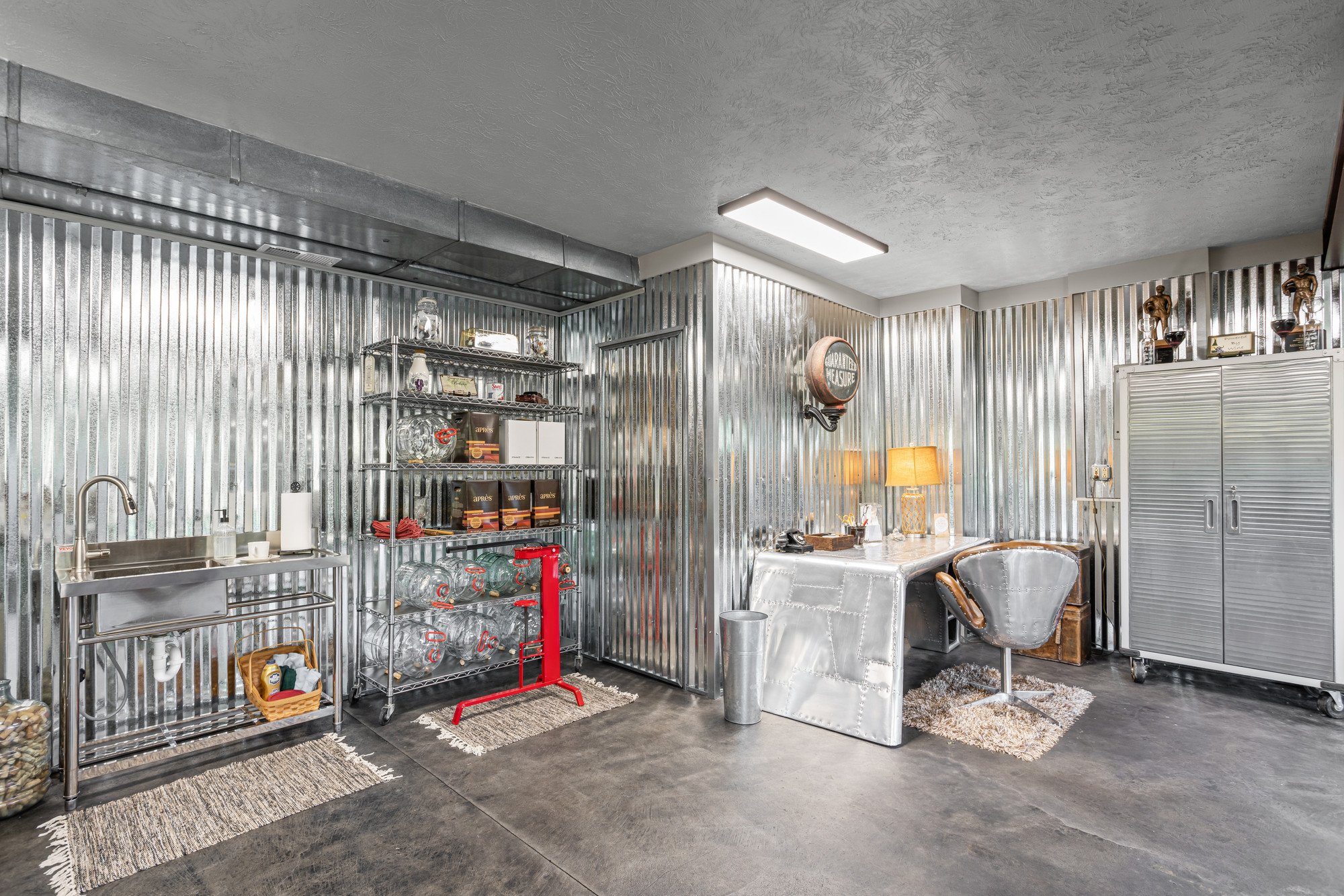 Industrial kitchen or workspace with metal walls, a sink, shelves with jars and boxes, a vintage metal table, a lamp, chairs, and storage cabinets.