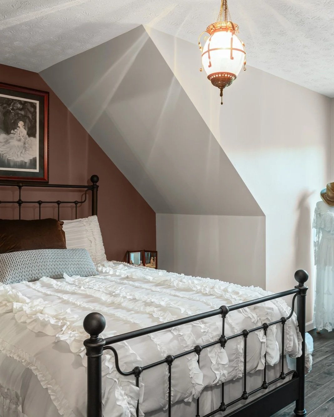 A bedroom with a metal bed, white ruffled bedding, decorative pillows, and a brown wall on the left side. Hanging from the ceiling is a vintage-style pendant light. There is a framed picture on the brown wall and a small side table. The room has an angled ceiling with a two-tone paint scheme, and natural light coming in from the right side.