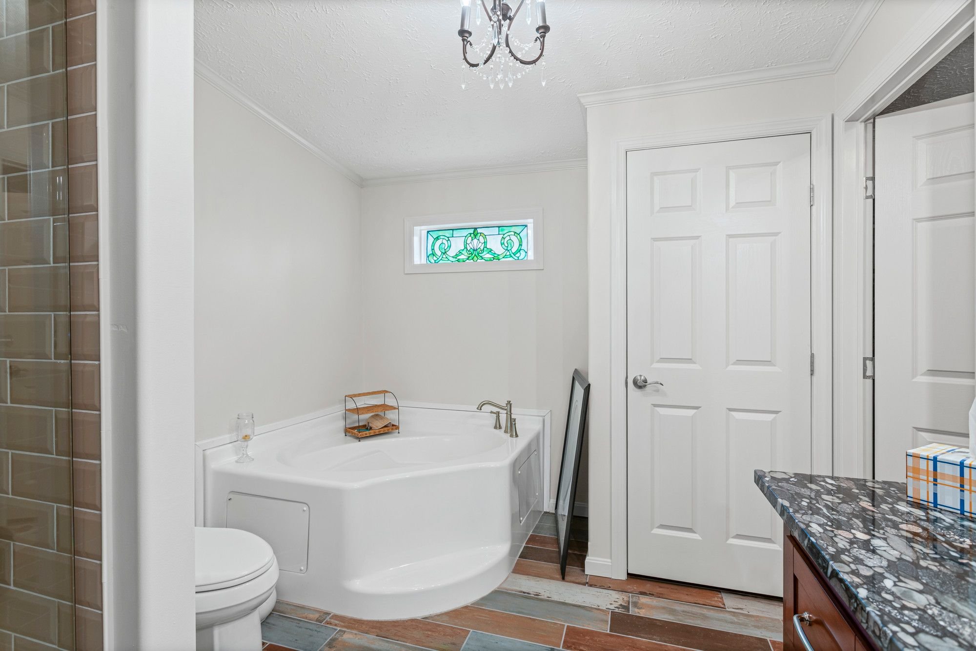 Bathroom with a bathtub, toilet, and granite countertop. Small stained glass window, white walls, textured ceiling, and a chandelier. Brown tile flooring.