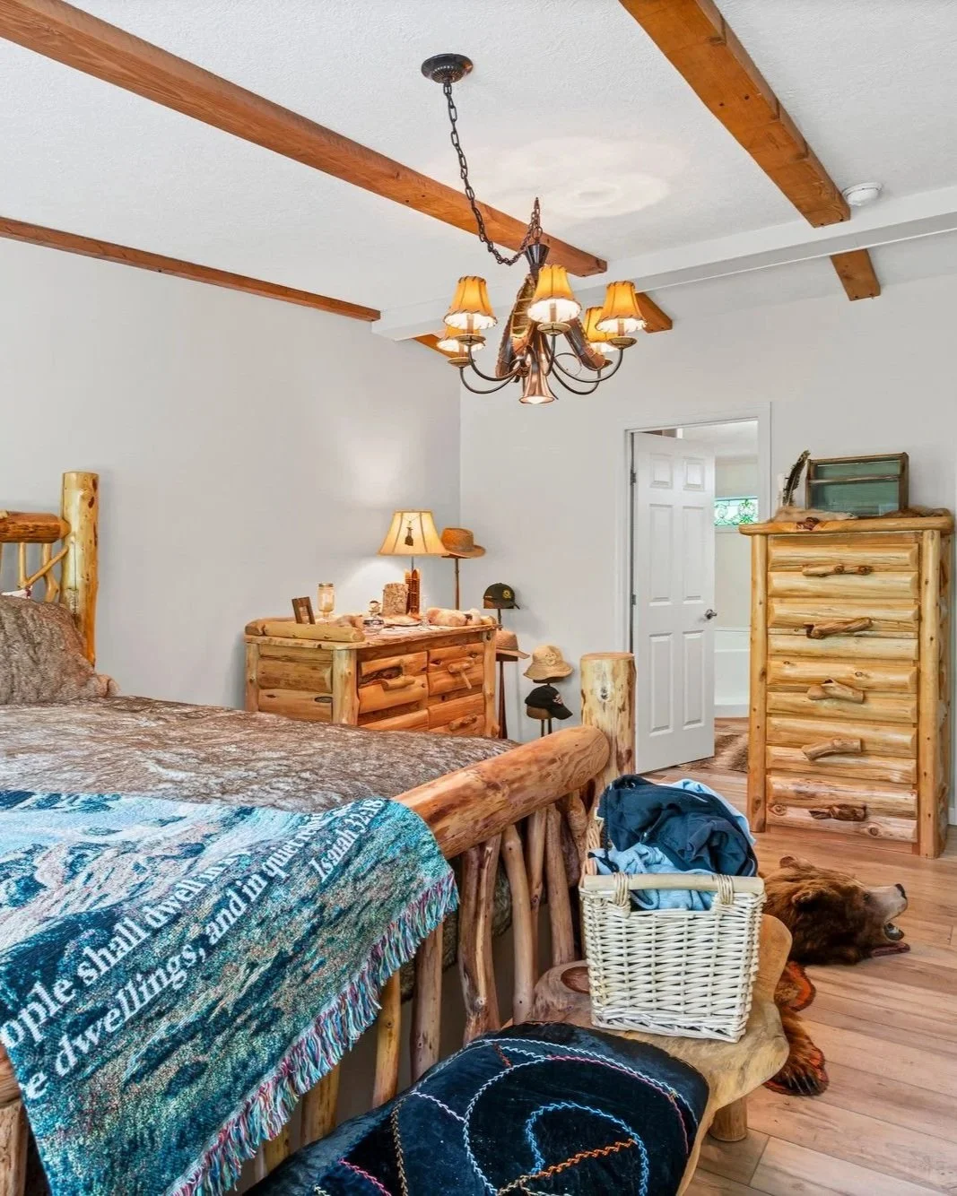 A rustic bedroom with log furniture, including a bed, dresser, and chest of drawers. There is a chandelier hanging from a ceiling with wooden beams, a lamp on the dresser, and hats on the floor. A dog is lying on a rug near the foot of the bed.