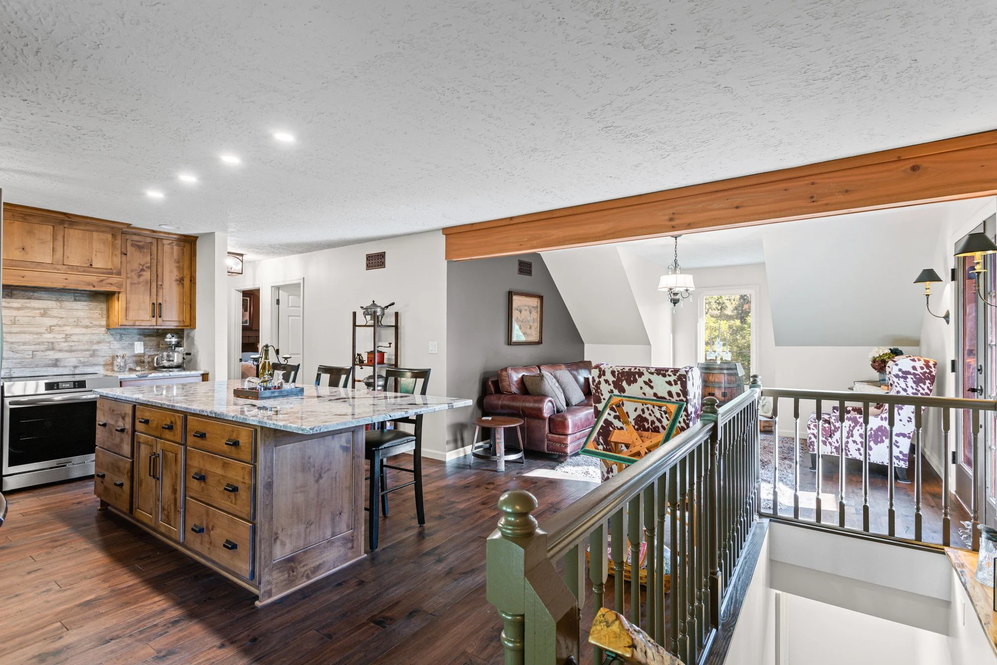 A spacious open-concept living room and kitchen with hardwood floors, wooden cabinets, and a large kitchen island with a granite countertop. The living area has a leather sofa, cow-print chairs, a hanging light fixture, and large windows letting in natural light. Part of a staircase with a green banister is visible in the foreground.