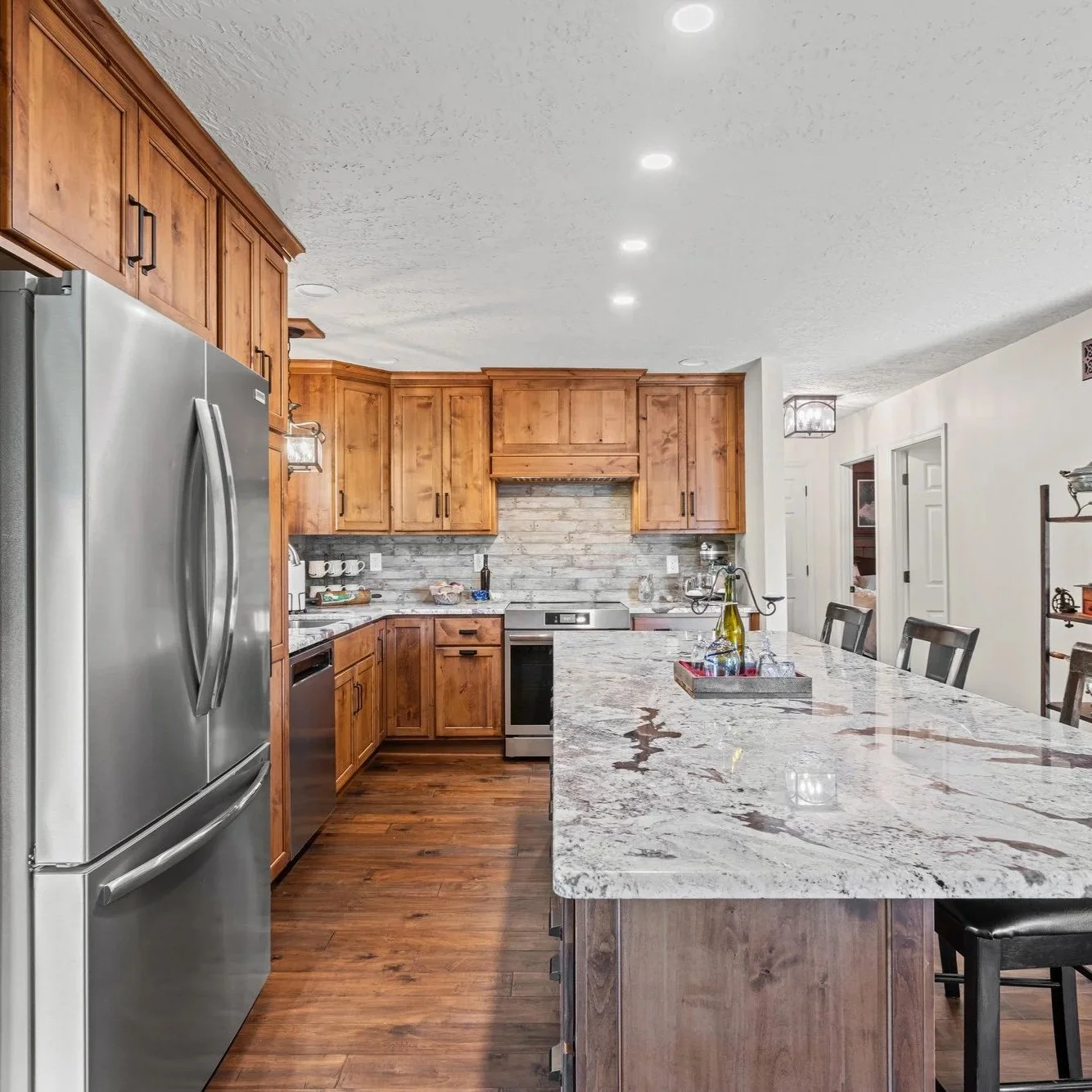 A modern kitchen with wooden cabinets, stainless steel refrigerator, and a large marble island with seating. The back wall features a stove, and there are various kitchen items on the countertops. The ceiling has recessed lighting, and there's a doorway leading to another room.