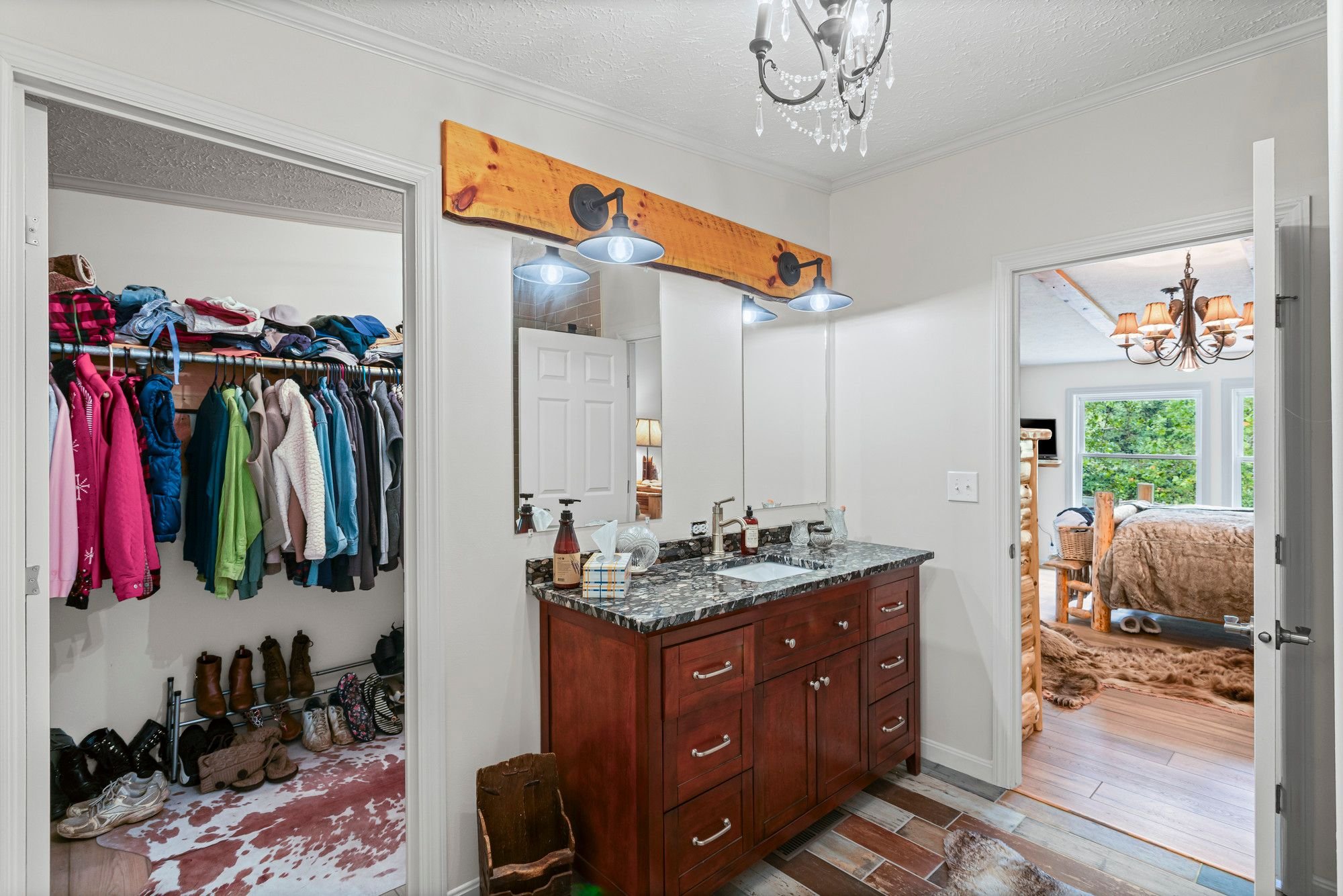 A bathroom with a granite countertop vanity, a mirror, and bright lighting. Adjacent to the bathroom is a walk-in closet with clothes and shoes, and a bedroom with a bed, a window, and a chandelier. The space has white walls and wood floors.