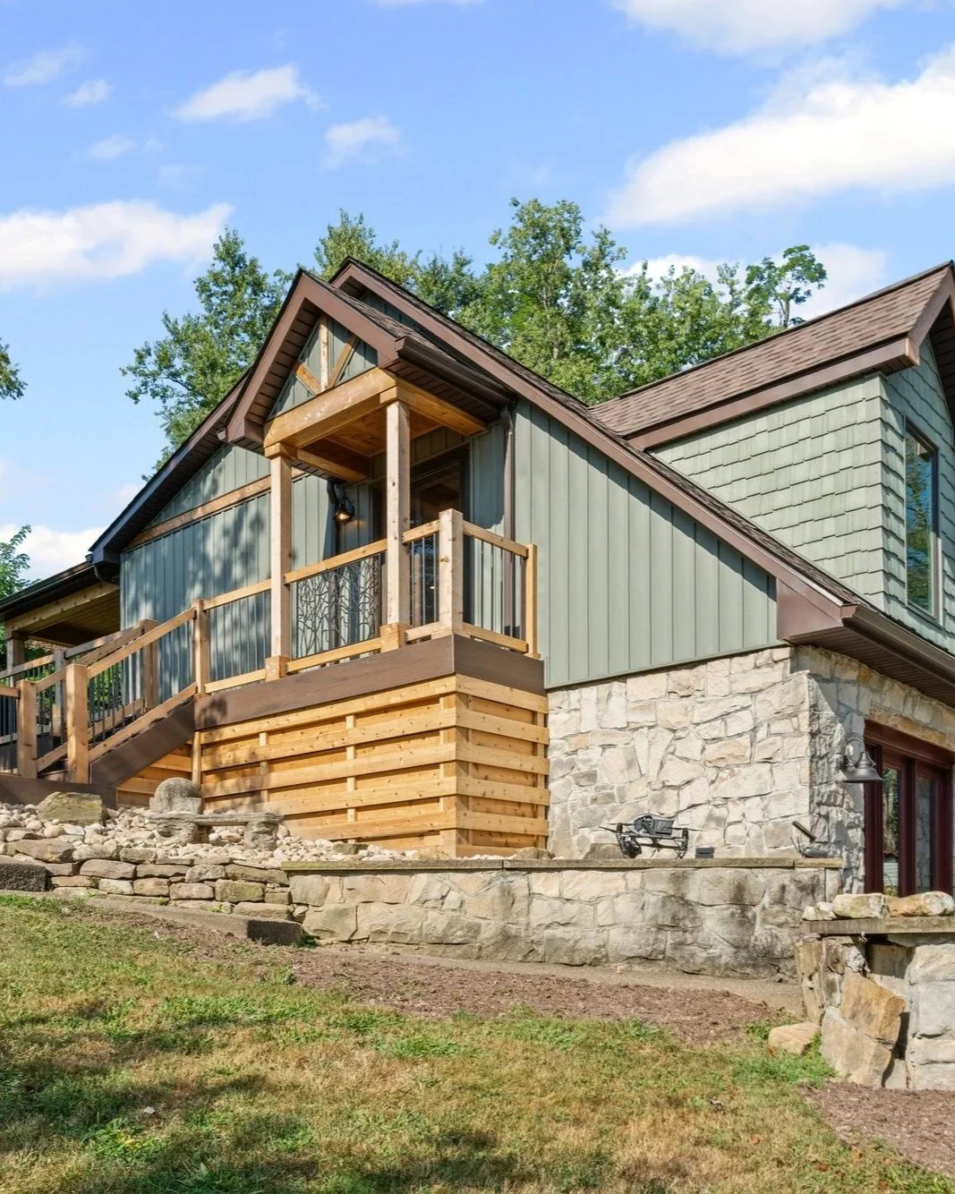 A house with a stone foundation, green siding, and a wooden balcony under a blue sky with white clouds.