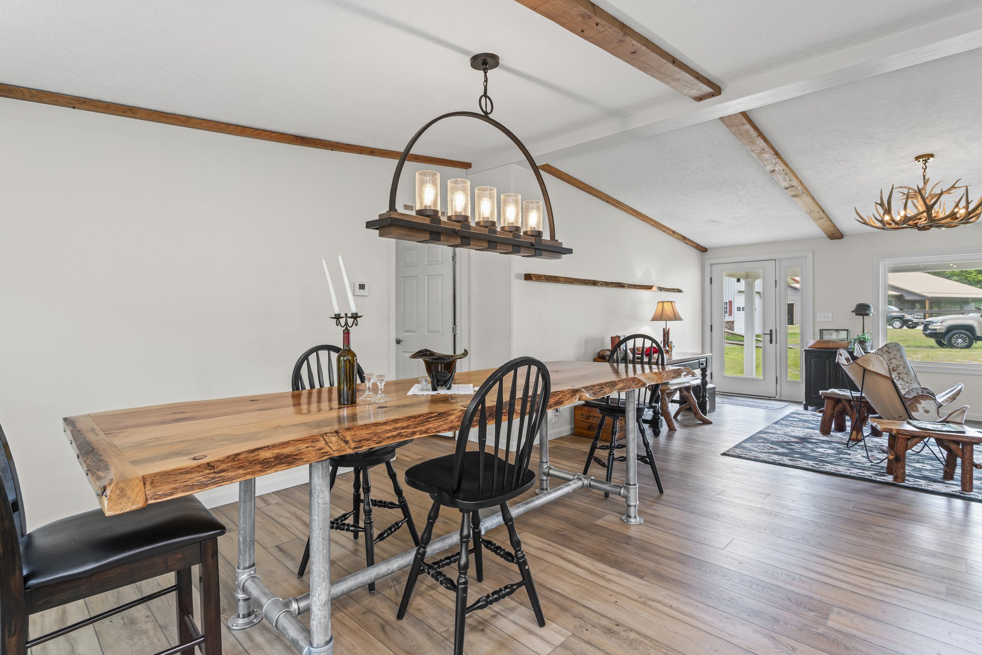 A dining area with a rustic wooden table, black chairs, and modern lighting fixtures hanging from the ceiling. Large windows and an open door show the outdoors. The ceiling has wooden beams, and there's a side area with a lamp, a chair, and various decorative items.