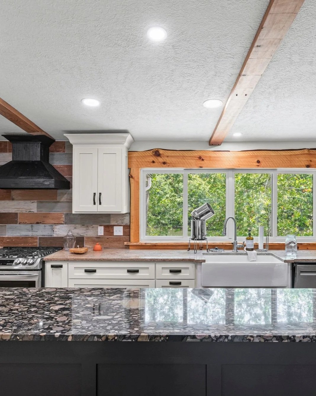 Modern kitchen with white cabinets, a granite countertop, a black range hood, and large window with a tree view.