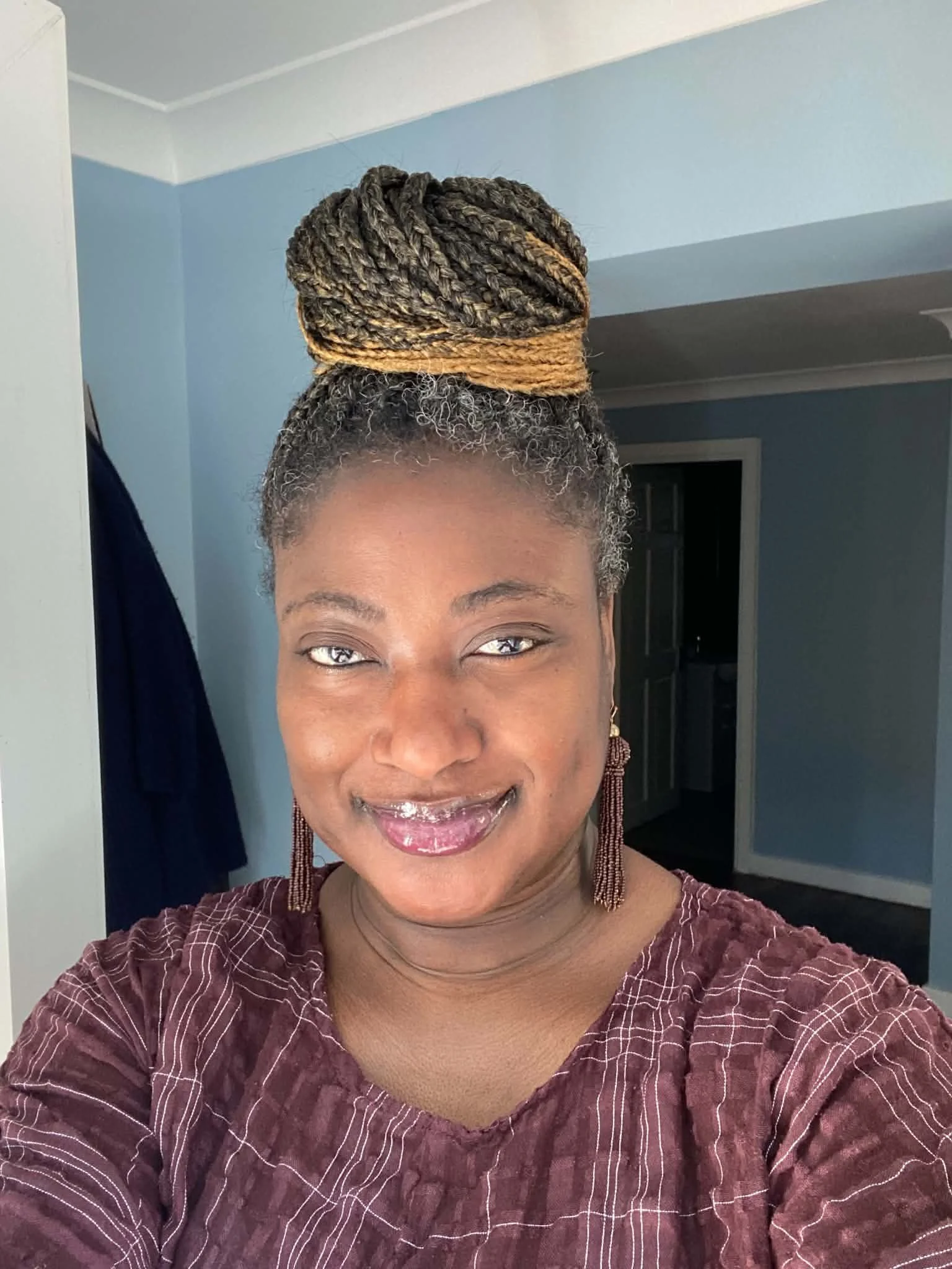 Close-up of a woman with styled multicolored dreadlocks, wearing earrings and a patterned maroon shirt, smiling in a room with light blue walls and a doorway in the background.