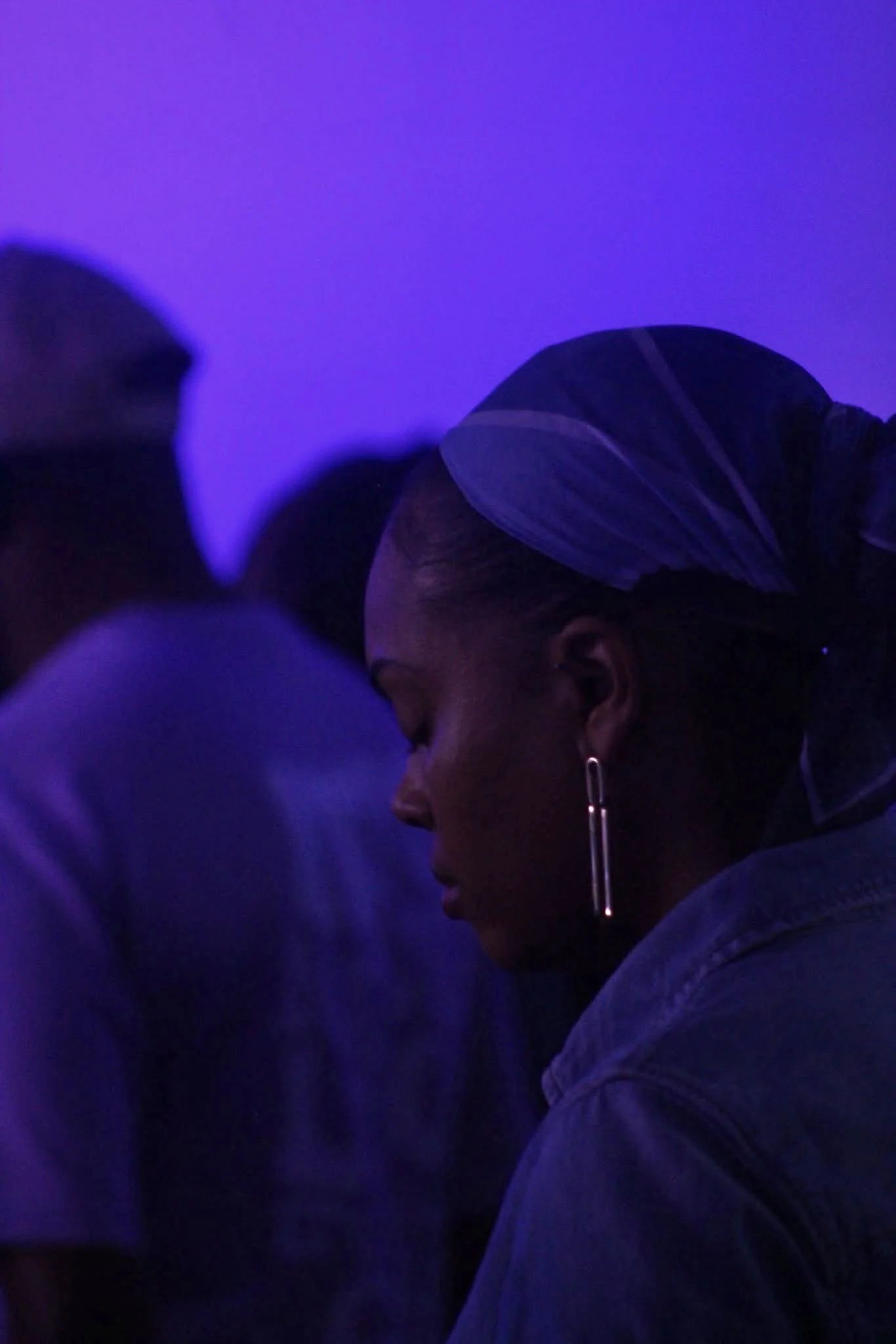 A woman with dark hair styled in a bun, wearing a striped headscarf and a jean shirt, is seen in profile against a purple-lit background.