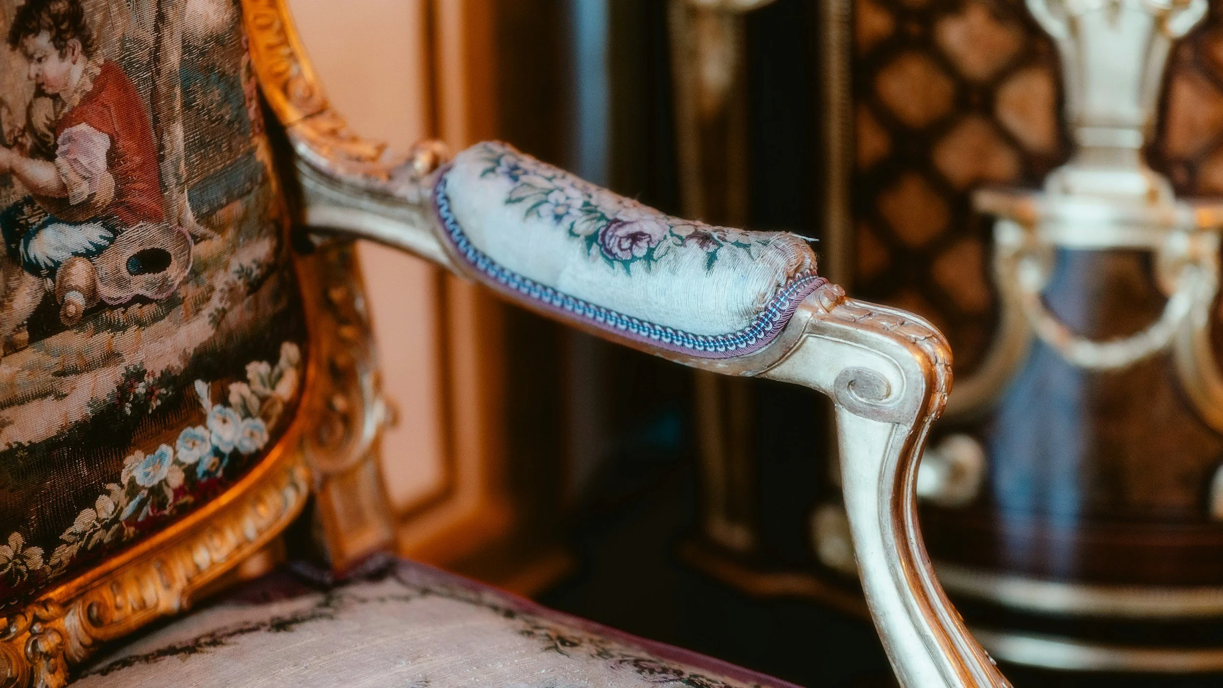 Close-up of an ornate antique chair with carved wood frame and floral upholstery, positioned in front of other similarly styled chairs.