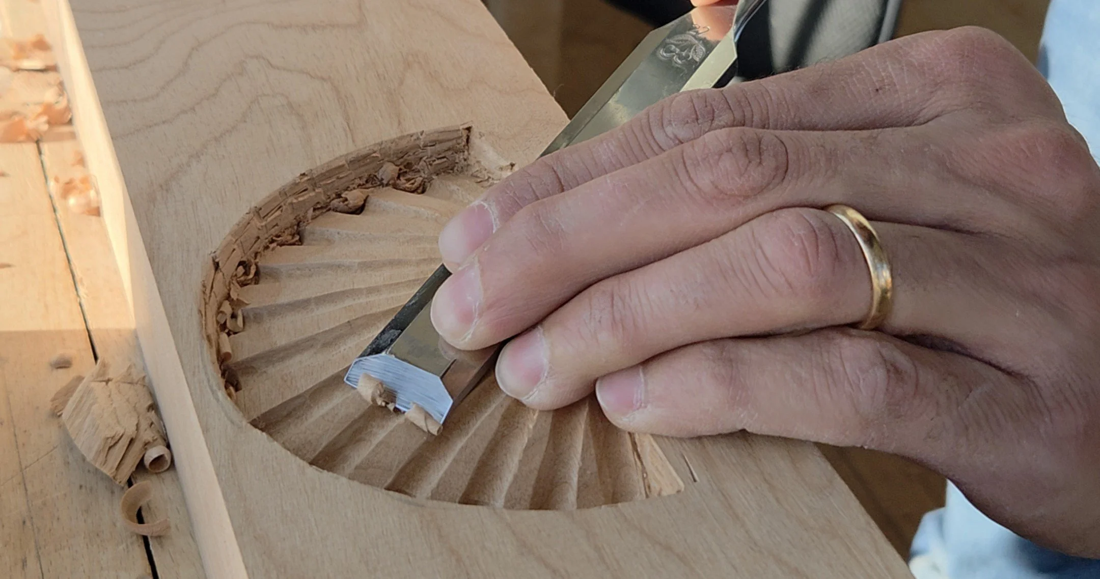 Close-up of a person's hand carving intricate details into a piece of wood using a chisel, with wood shavings around the work area.