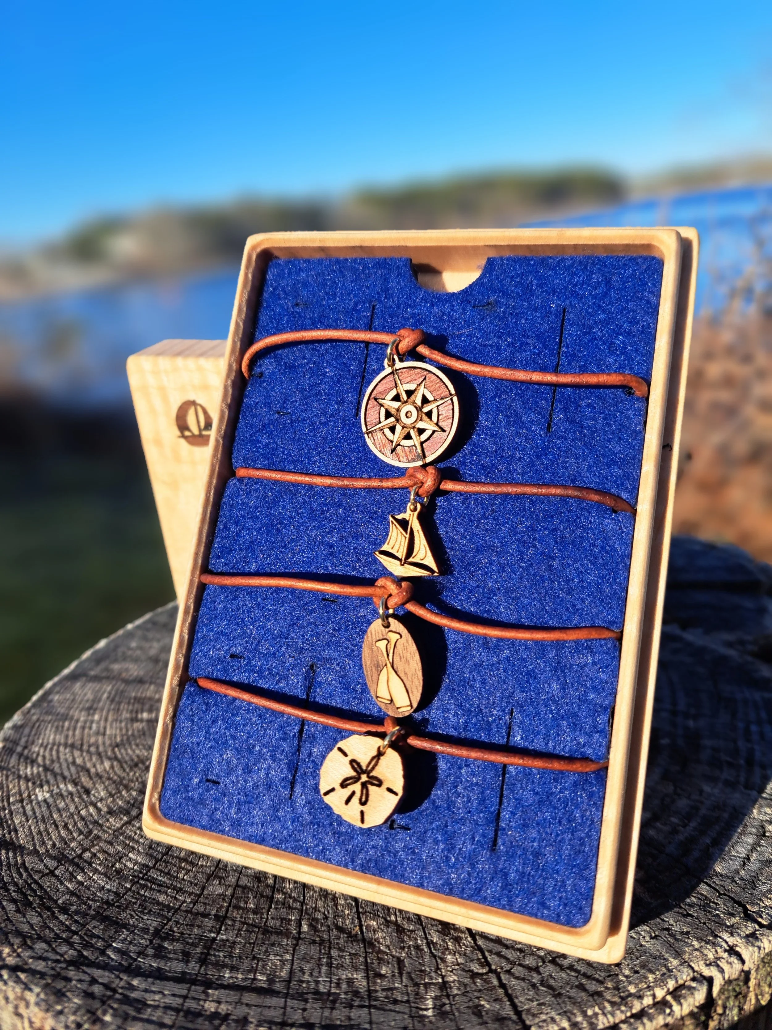 A wooden box with a blue velvet interior displays wooden jewelry charms on leather string. The charms include a ship, a lighthouse, and a globe, set outdoors with water and trees in the background.
