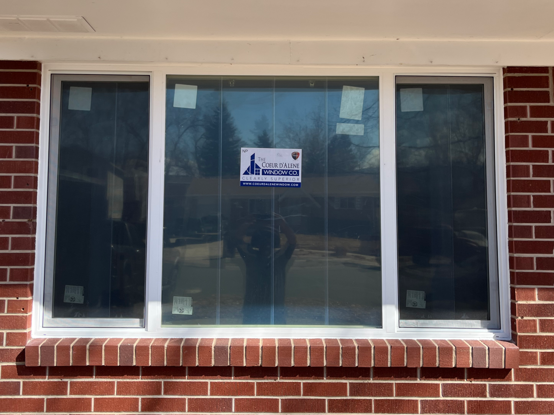 A large window with three glass panels set in a brick building exterior, with a sign on the window advertising a window company.