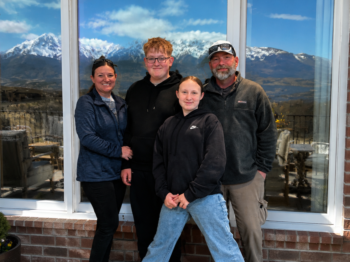 A family of four posing in front of a large window with snow-capped mountains in the background.