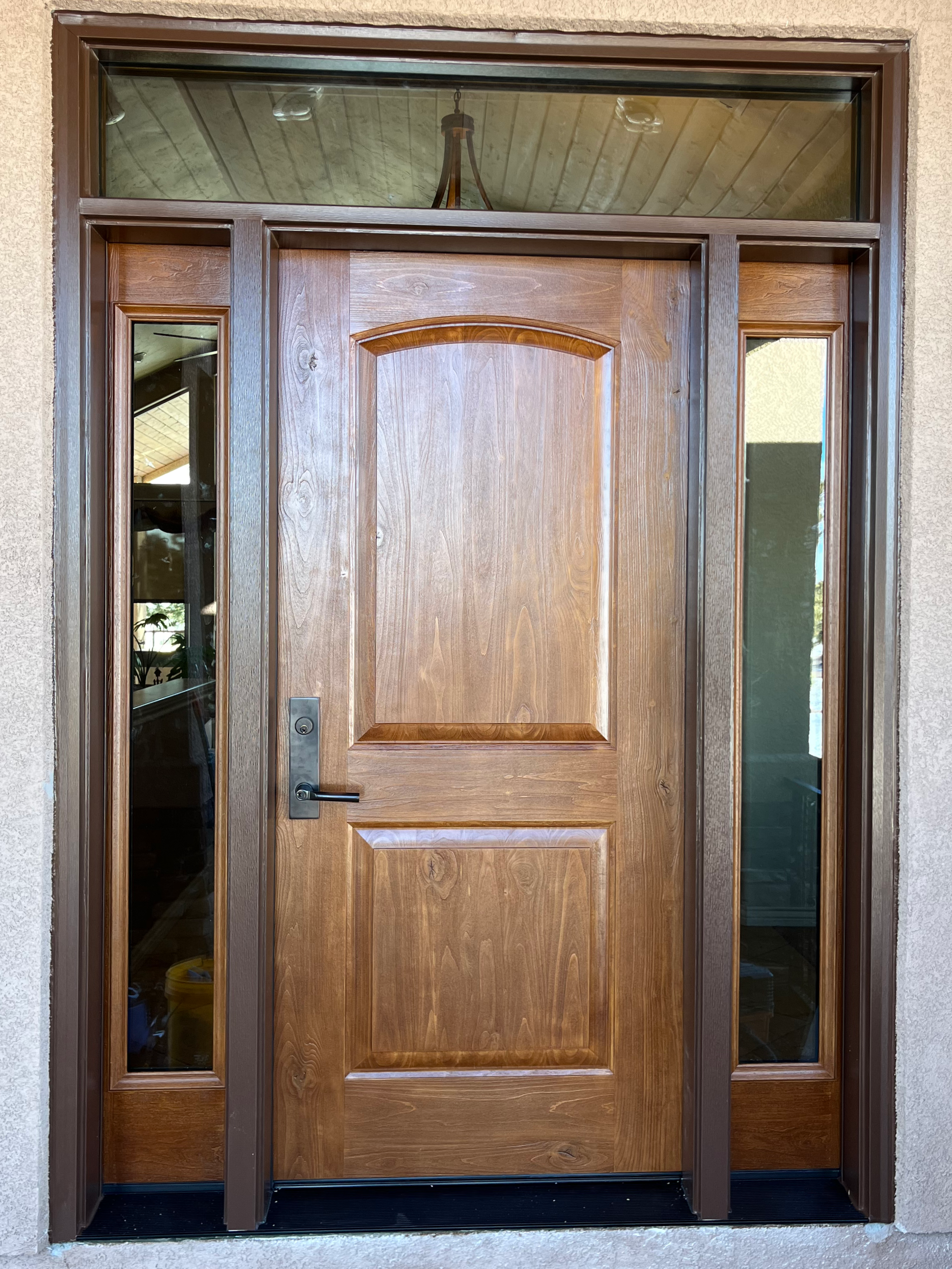Wooden front door with glass side panels and a transom window above, set in stucco exterior wall. A chandelier is visible through the transom window.