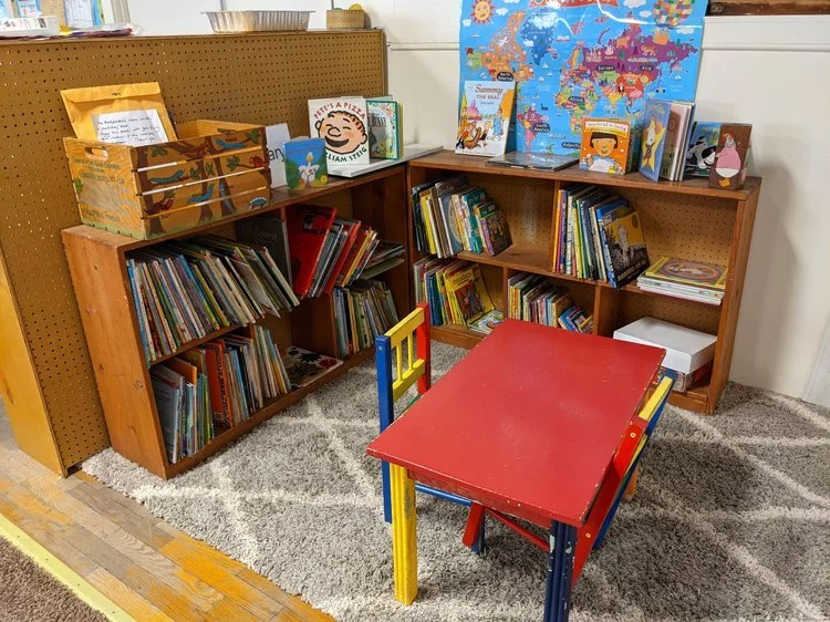 Children's area with wooden bookshelves filled with children's books, a small colorful table with chairs, a world map poster on the wall, and a yellow pegboard.