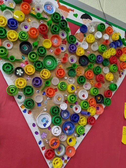 A wall display with colorful plastic bottle caps and small decorative items arranged in the shape of a heart.
