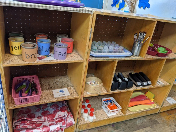 Organized wooden shelves with craft supplies, including paint cups labeled 'yellow', 'blue', 'purple', and 'red', a pink basket with headbands, a container of glue sticks, scissors, markers, and various art materials.