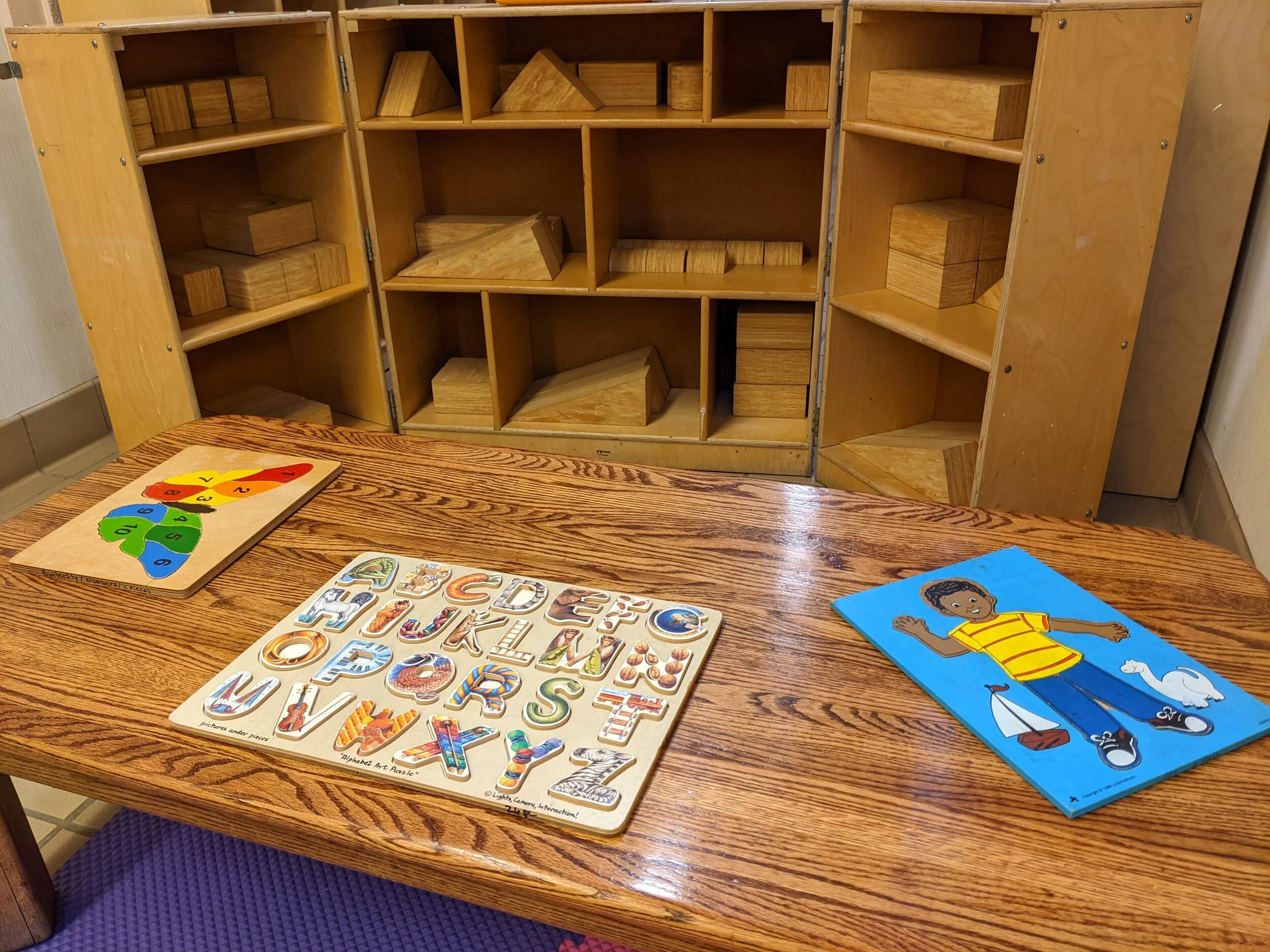 Wooden shelf with various wooden geometric shapes and blocks, and educational children's puzzles and cards on a wooden table in the foreground.