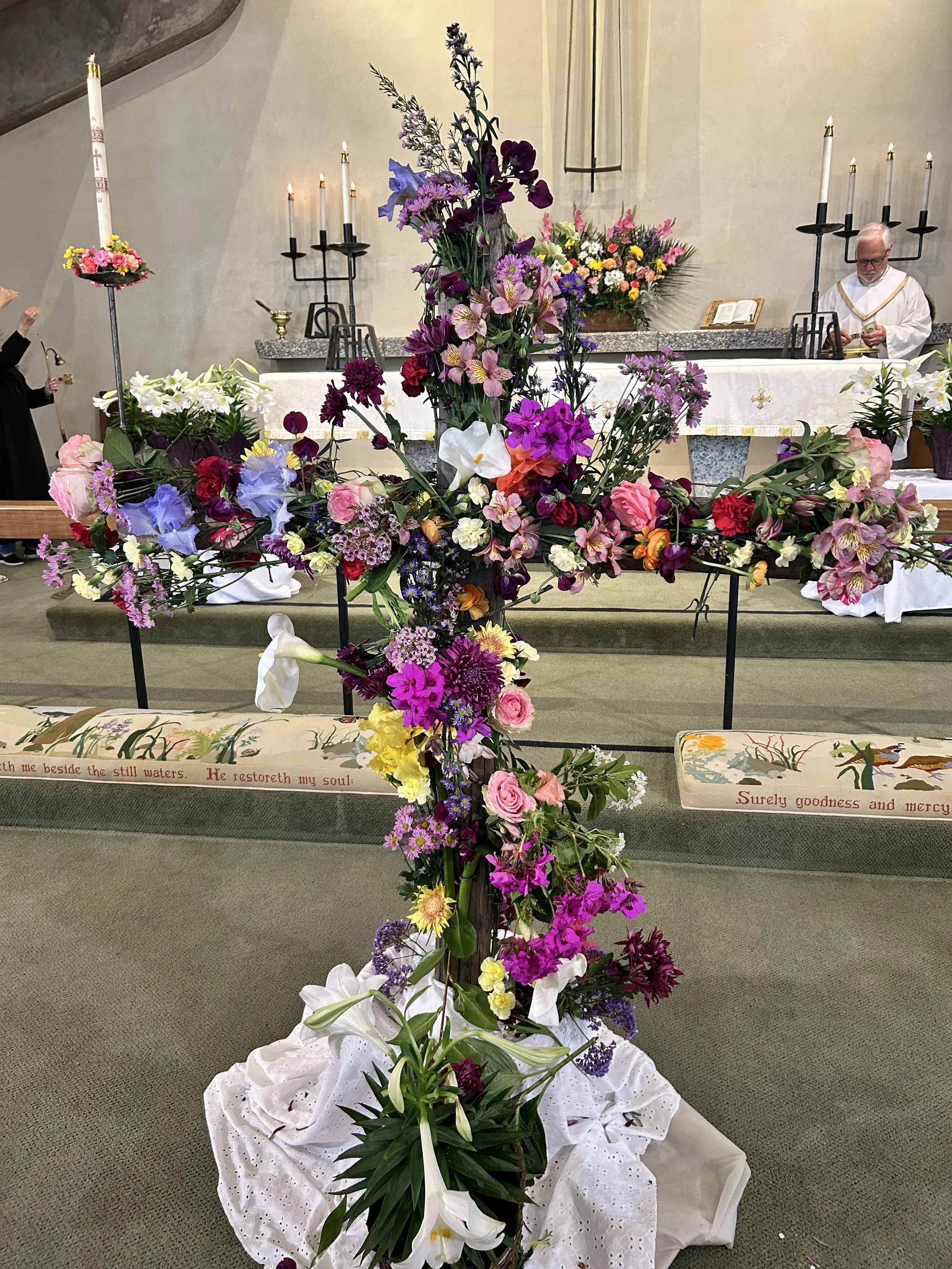 Large floral arrangement with pink, purple, white, and yellow flowers, set in front of a church altar decorated with candles and more flowers. A minister is in the background preparing for a service.
