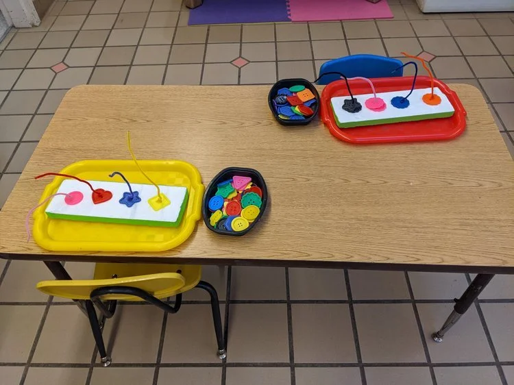 Table with two sorting trays of buttons and pins for a children's threading activity and a small black container of colorful buttons and shapes.