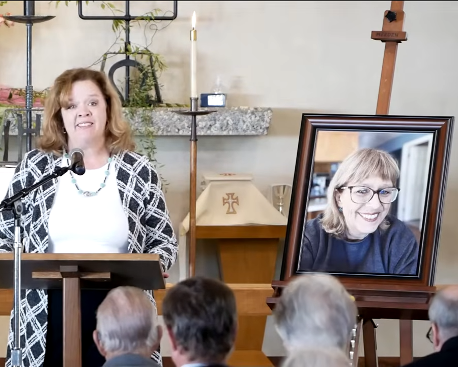 A woman giving a speech at a lectern during a memorial service, with a framed photograph of another woman on an easel nearby.