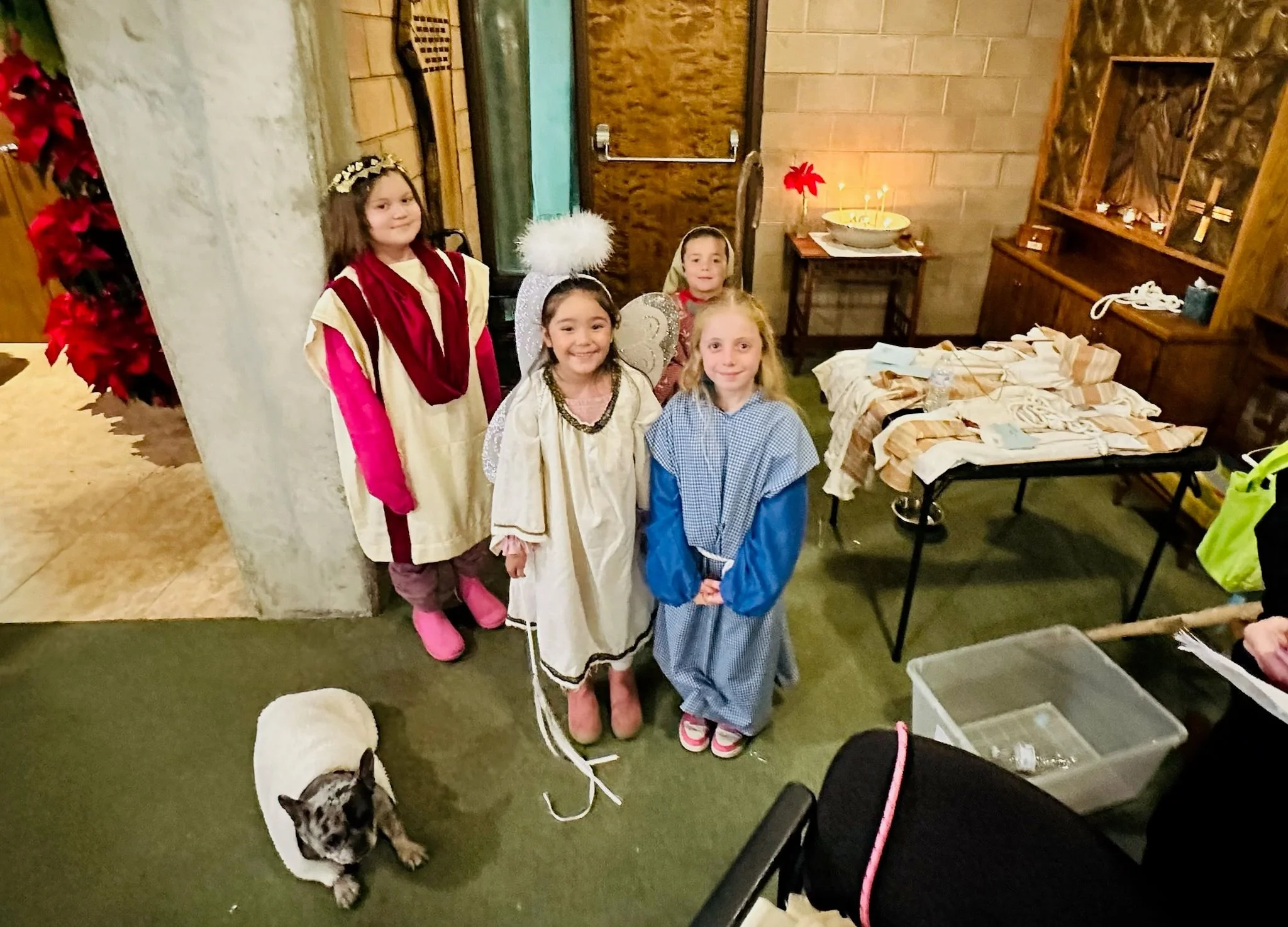 Five children dressed in costumes, including angels and a biblical character, are standing indoors near a table with wrapped gifts and a small black and white dog. The background includes a wooden door, a side table with a candle, and a wooden display shelf.