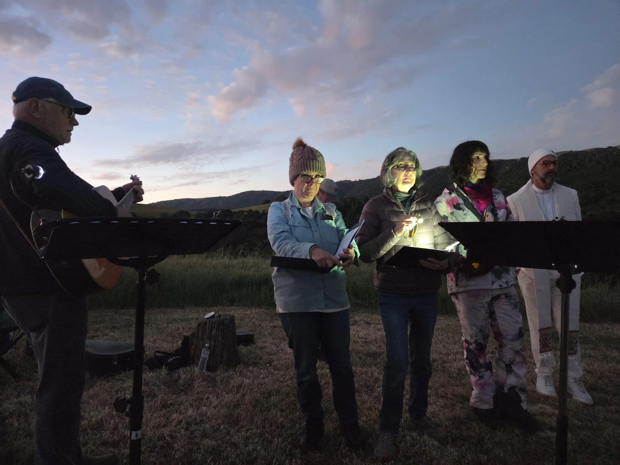 A group of five people standing outdoors during dusk, with mountains in the background, some holding flashlights or reading materials, and a musician playing a guitar on the left.