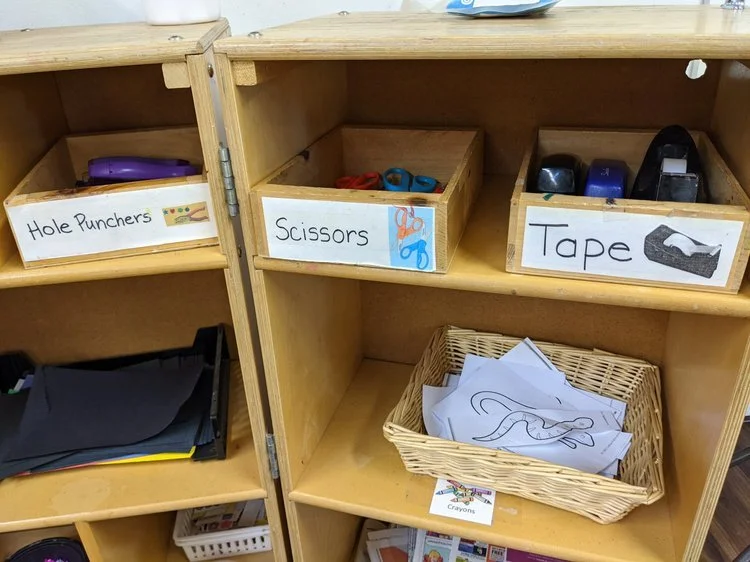 A wooden storage cabinet with labels for hole punchers, scissors, and tape, containing office supplies and papers.