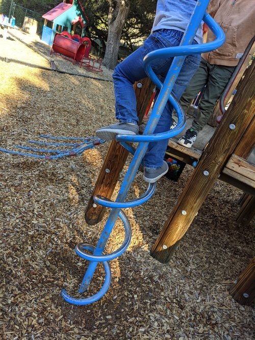 Child climbing on a blue spiral-shaped metal playground slide at a park, with other playground equipment and trees in the background.