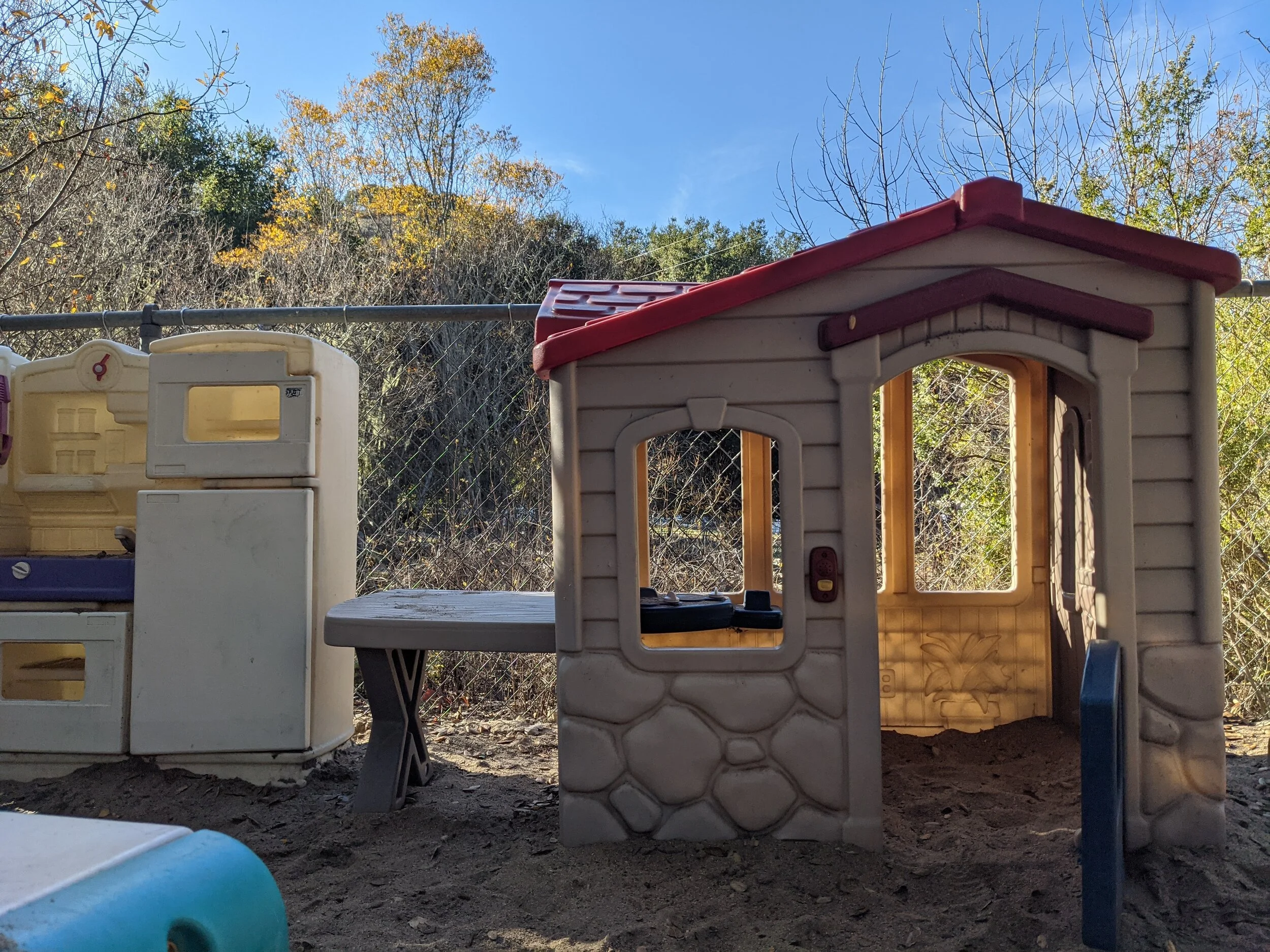 A playground set with small plastic houses and a chair, outdoors near a fenced area with trees and a clear blue sky.