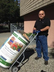 Man standing next to a large planter shaped like a roll of garden soil, holding a garden trowel.
