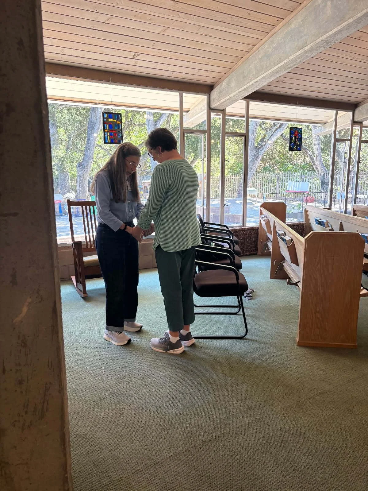 Two women holding hands and bowing in a room with large windows, chairs, and a church pew, with trees visible outside.