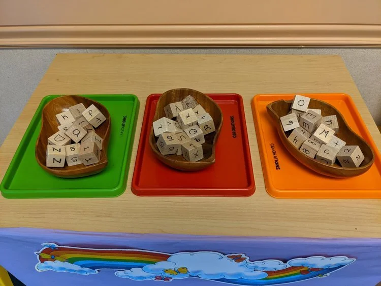 Three trays with wooden bowls containing lettered blocks on a table with a colorful rainbow and clouds decoration.